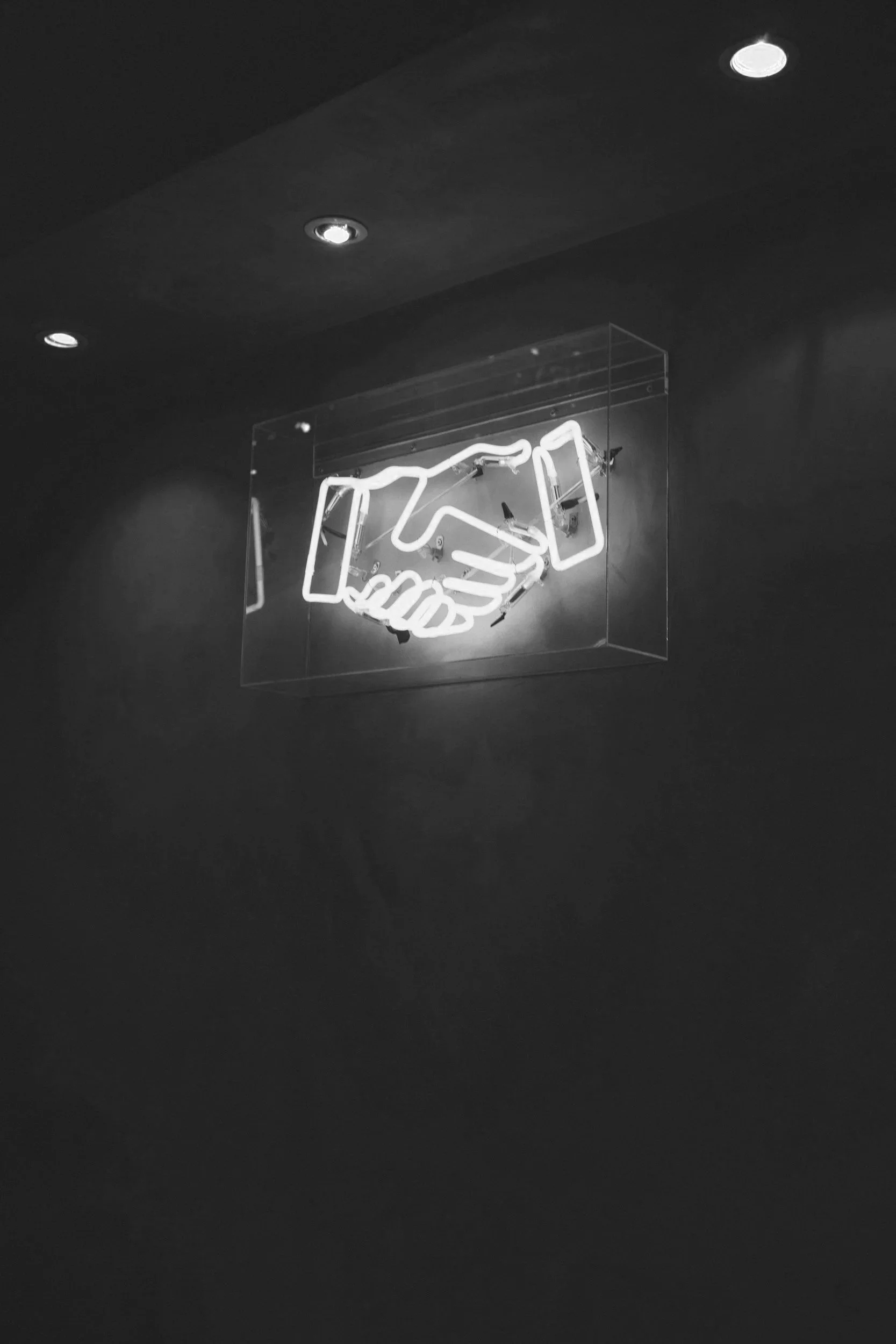 Neon sign of a handshake inside a transparent box on a dark wall ceiling with recessed lights.
