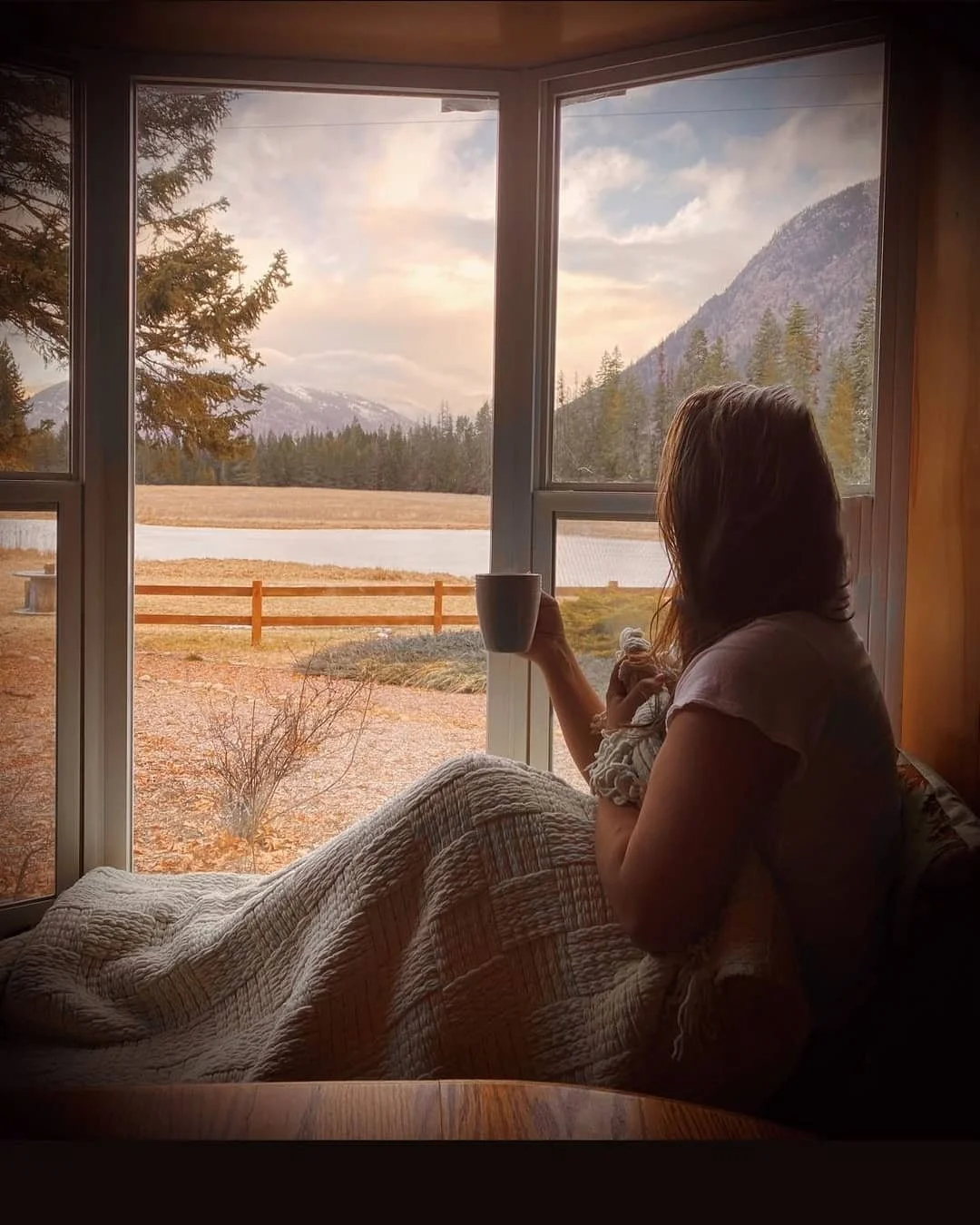 A woman wrapped in a blanket looking out at a lake and mountains at sunrise