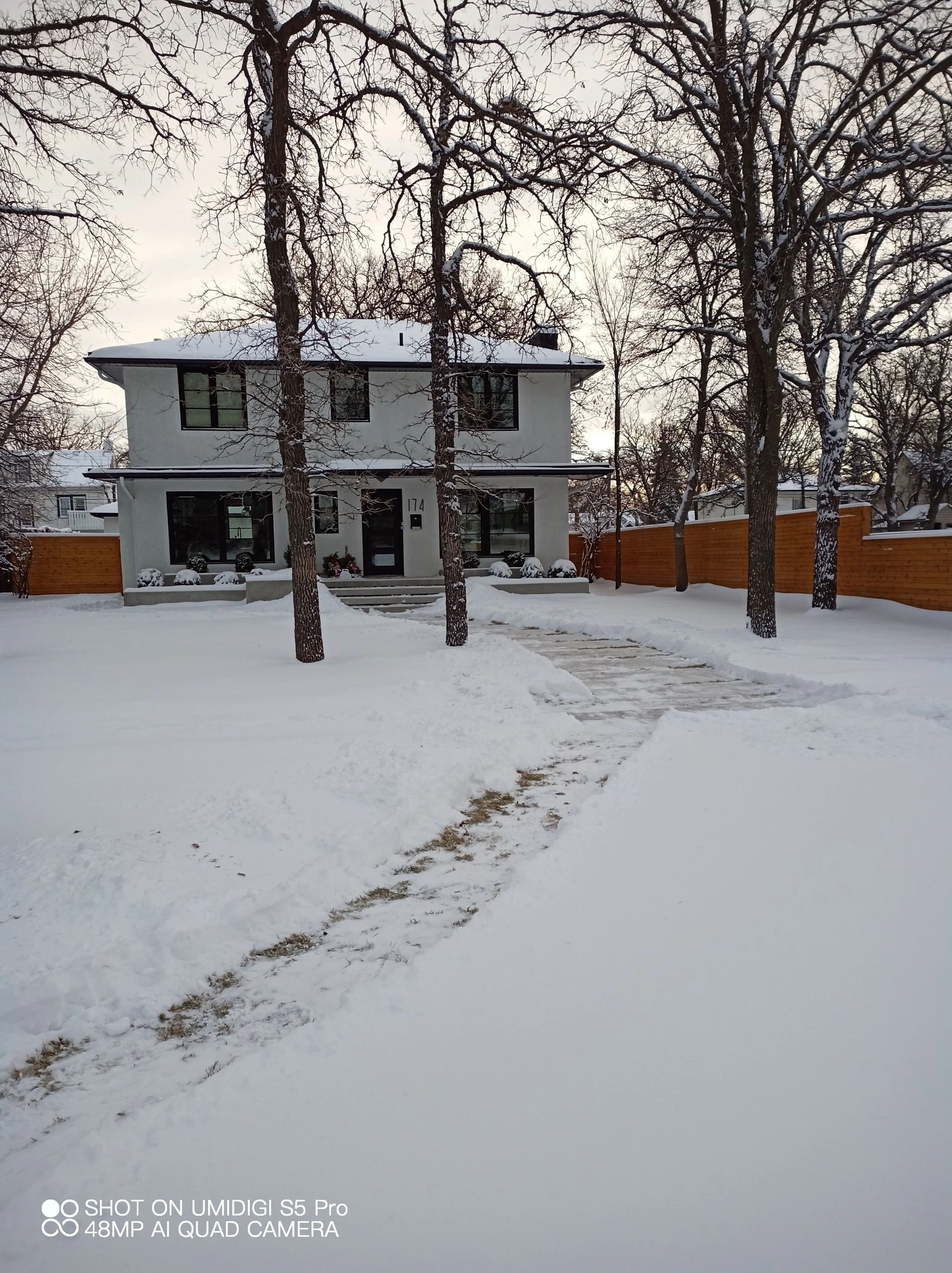A house covered in snow with a cleared pathway leading to the front steps. The house is white, has two stories, with black window frames, and a black door. There are several trees in the yard, some with snow on their branches, and a wooden fence on either side of the property.