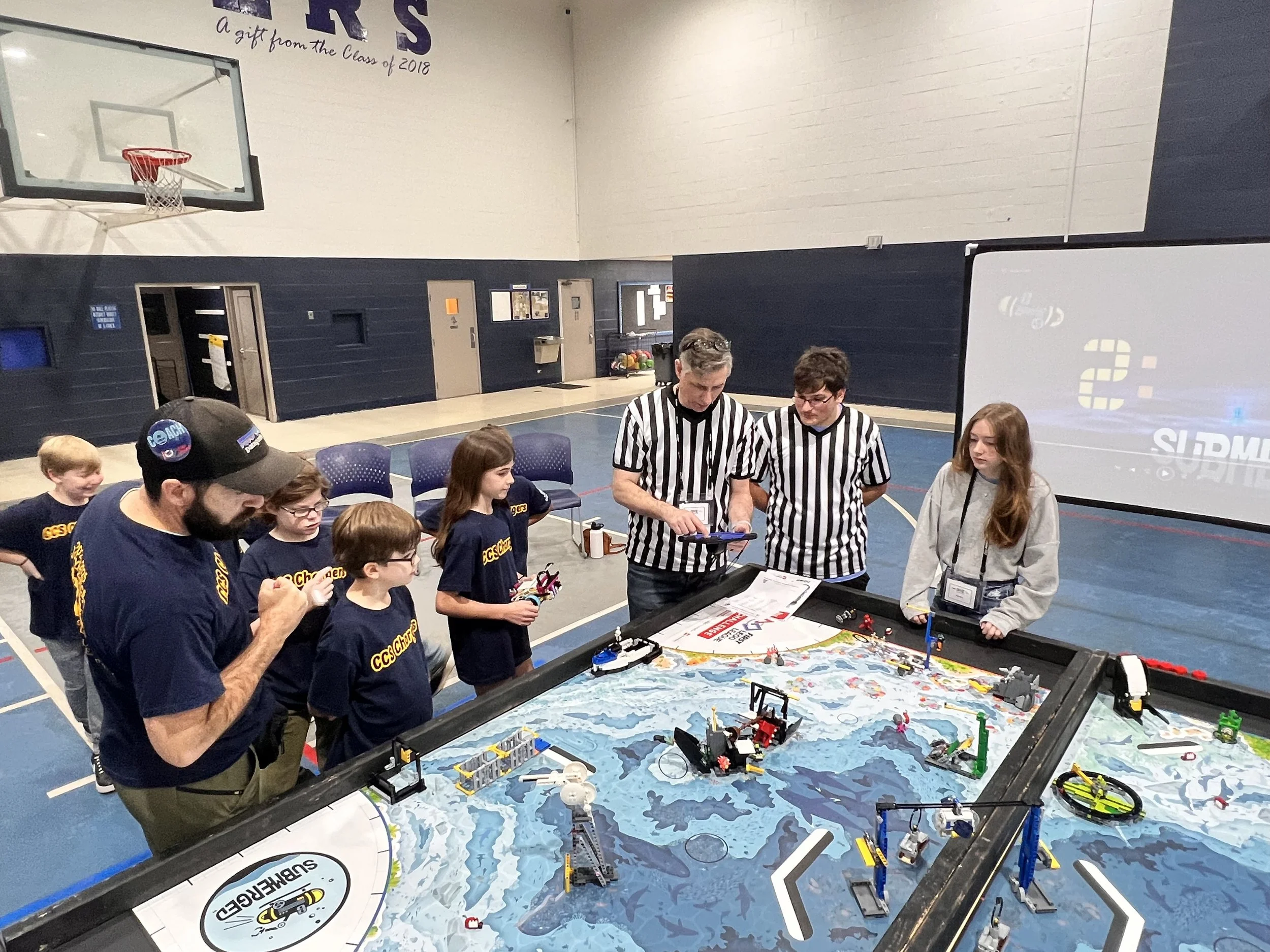 Children and adults gathered around a robotics competition table in a gymnasium, with kids wearing matching navy shirts and adults wearing referee shirts, participating in a robot challenge.