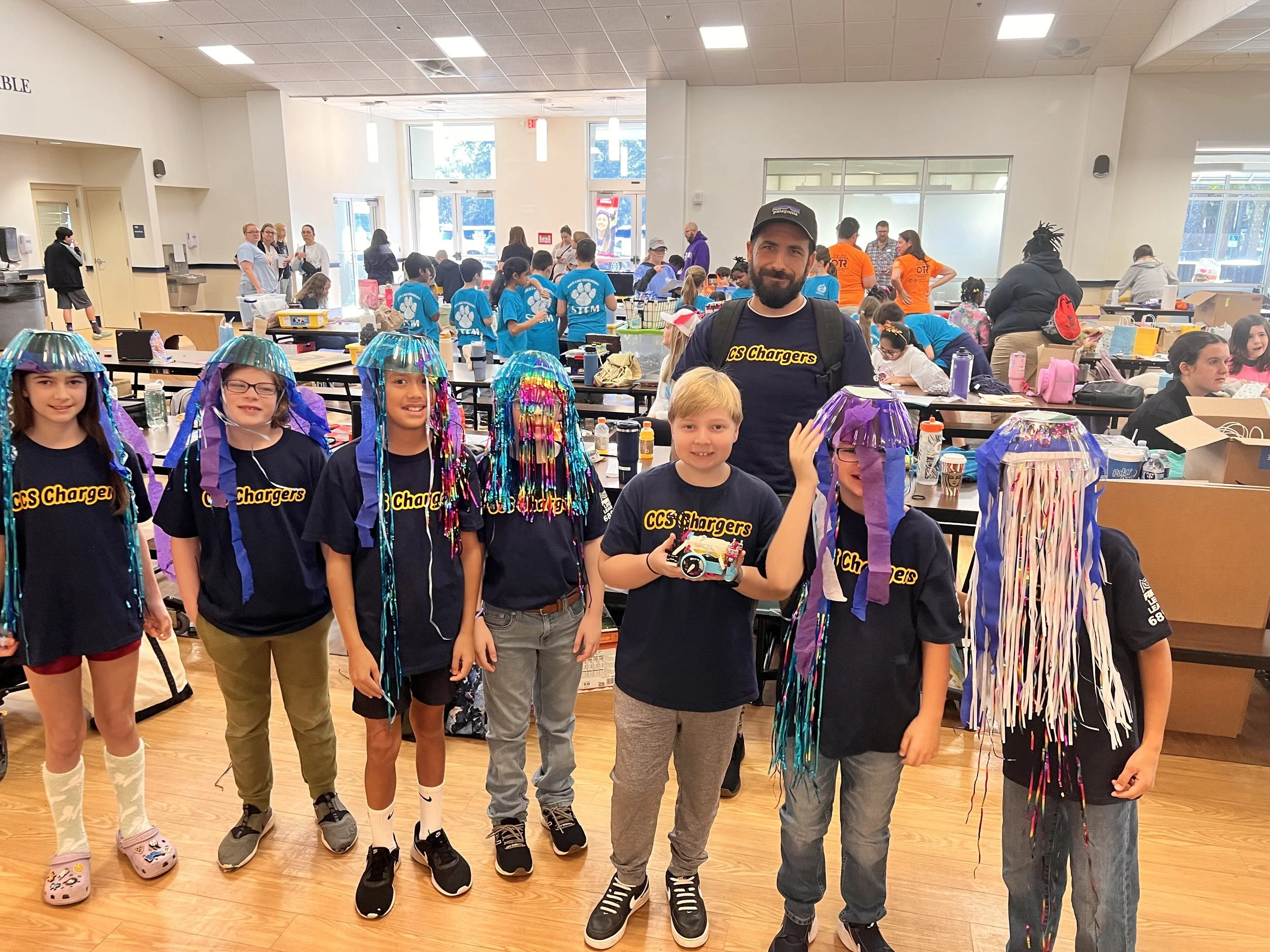 Children wearing CCS Chargers shirts, some with colorful streamers on their heads, standing with an adult in a room filled with other kids and adults.