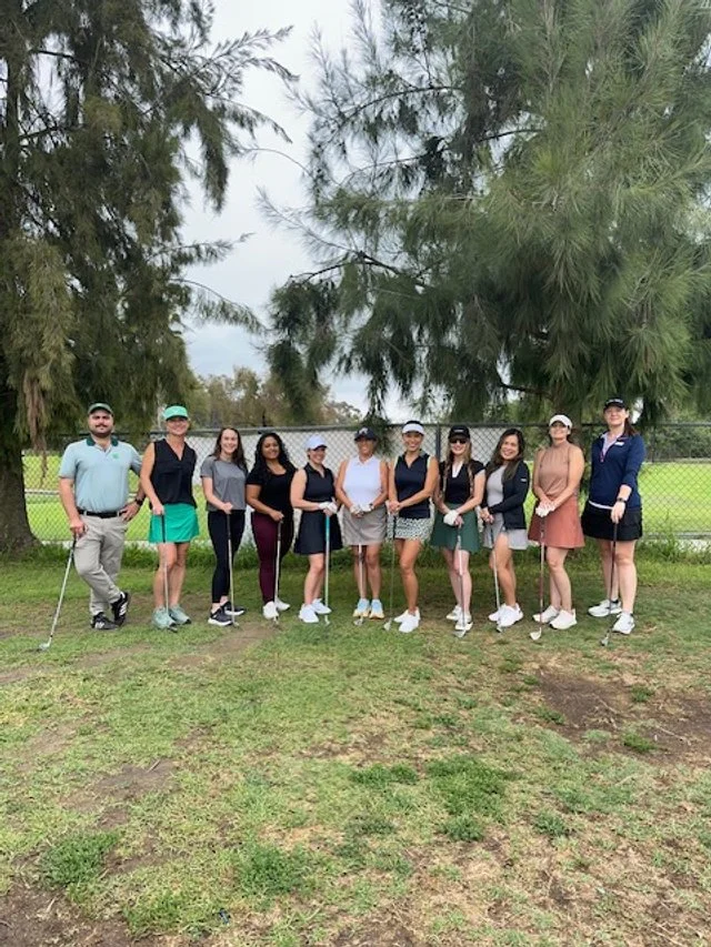 Group of eleven women and one man standing on grass at a golf course, holding golf clubs, with trees and a chain-link fence in the background.