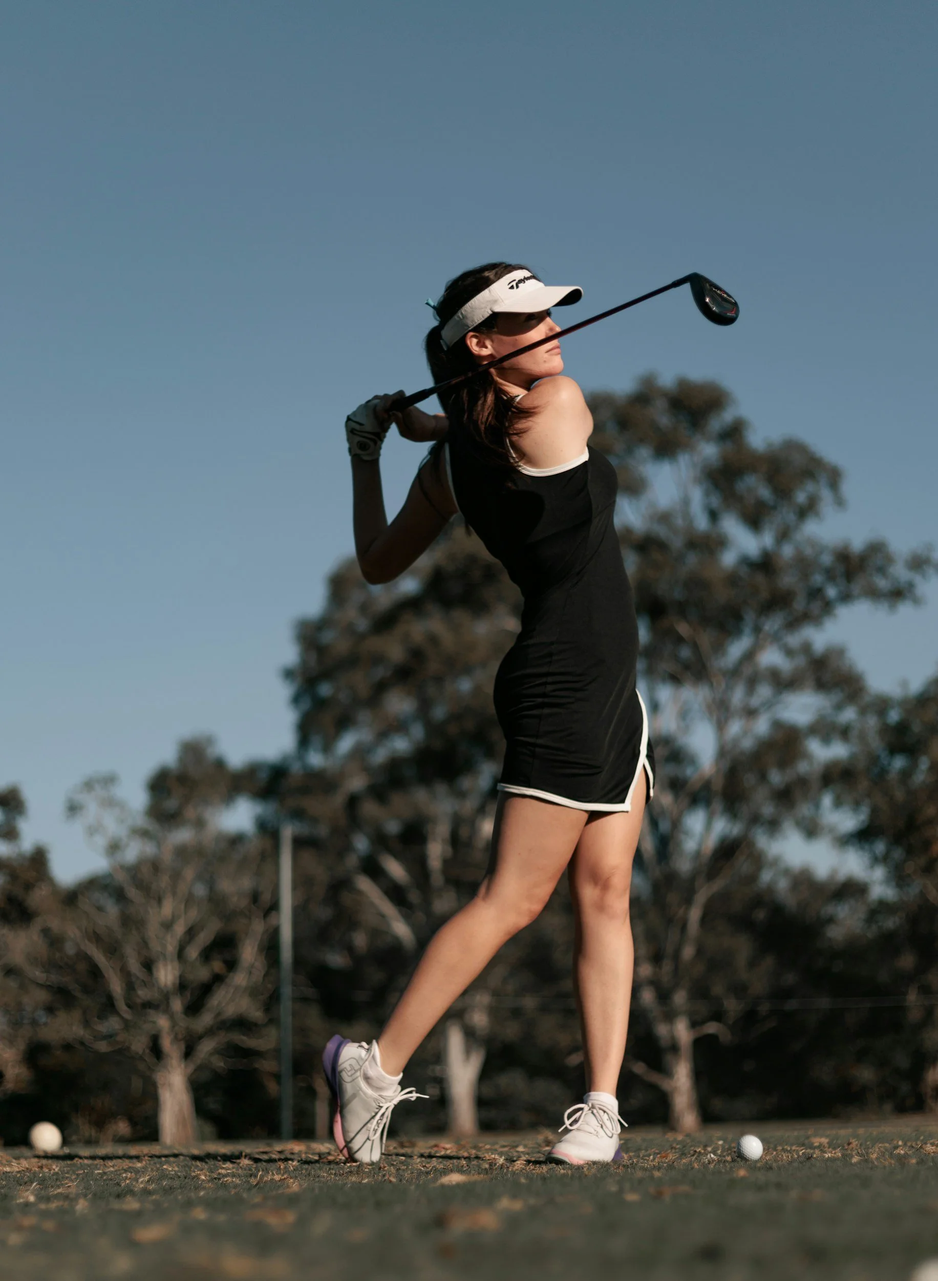 A woman playing golf on a field during late afternoon, swinging a golf club with a golf ball on the ground and trees in the background.