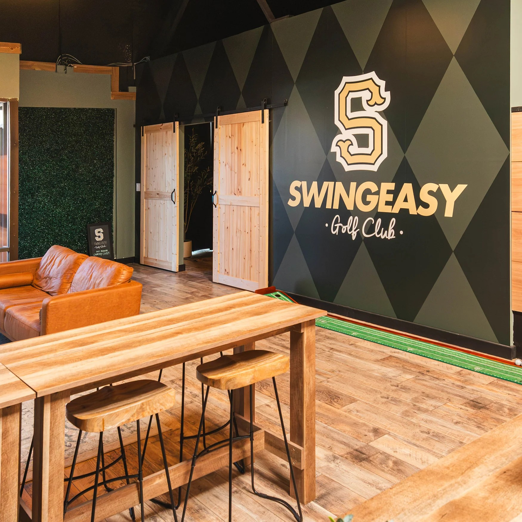 Interior of a golf club lounge with wooden tables, black chairs, a green hook-and-loop mat on the floor, a dark geometric accent wall with a large 'S' logo and text 'SWINGEASY Golf Club.' Wooden sliding doors and green foliage in the background.