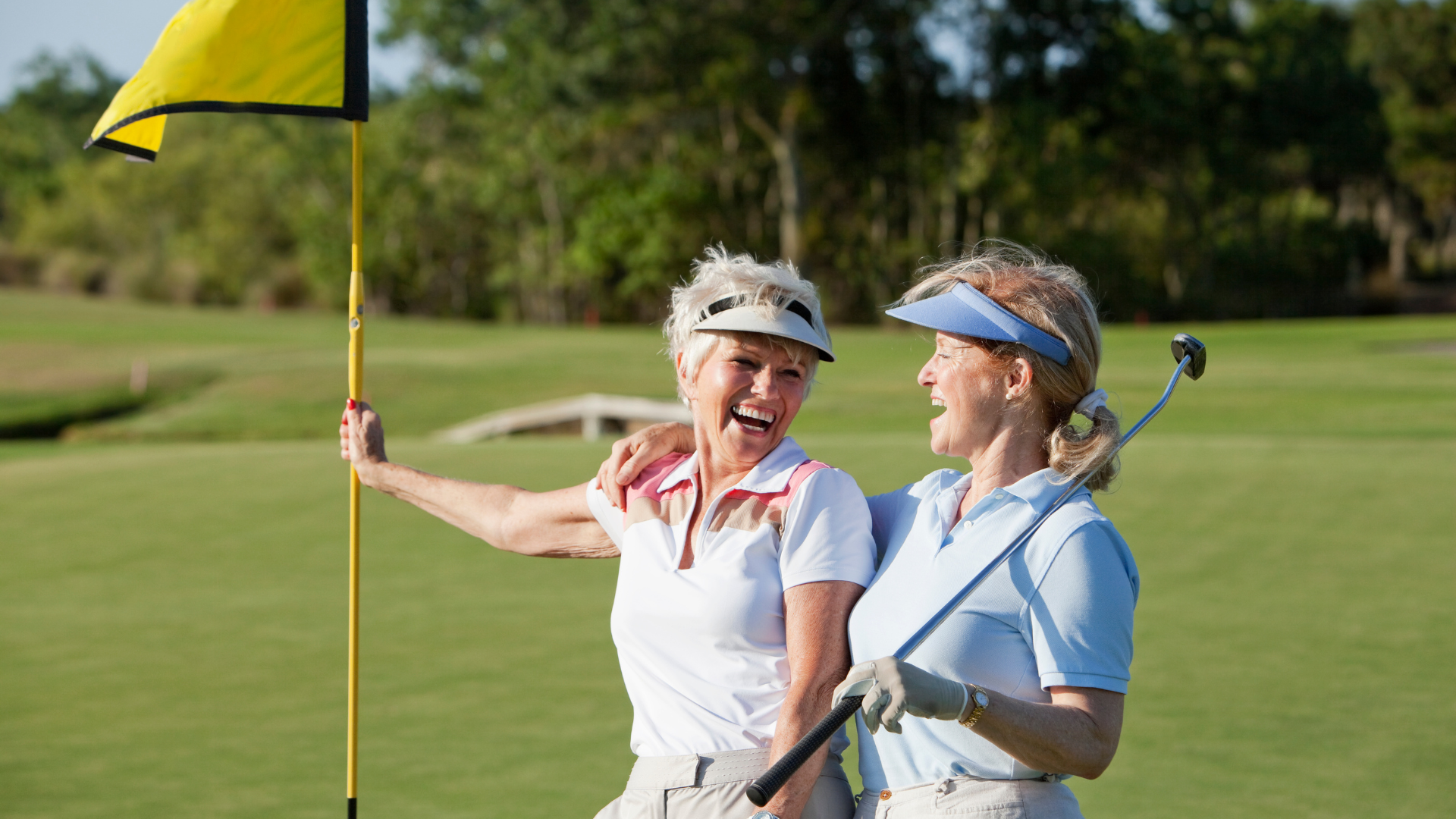 Two women celebrating on a golf course, one holding a golf flag, both wearing visors and smiling.