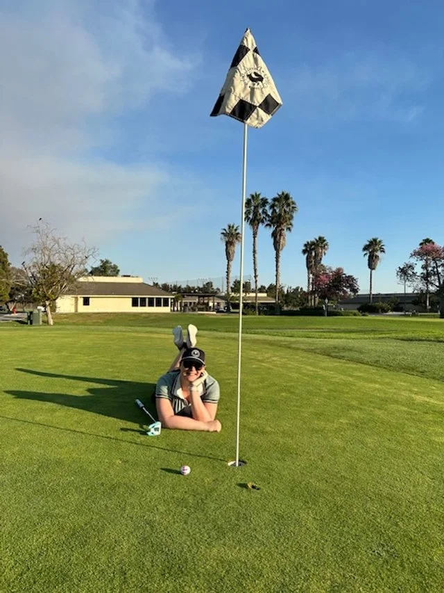 Person lying on the grass at a golf course, wearing bunny ears, sunglasses, and a cap, resting their chin on their hand near a golf ball and a putter, with a flags on the hole, palm trees, and a building in the background.