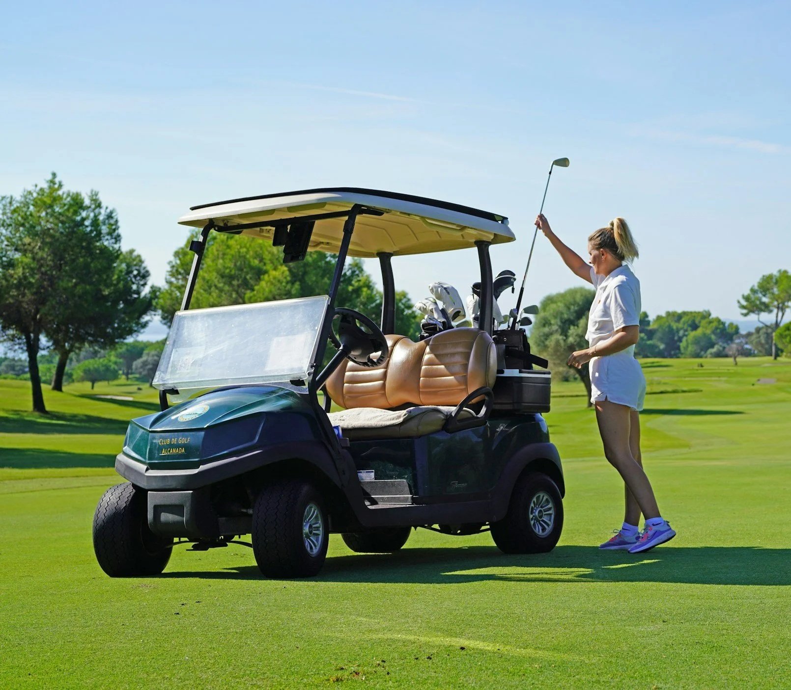 A woman in a white outfit and sneakers standing next to a black golf cart on a golf course, holding a golf club in one hand, with trees and grass in the background.