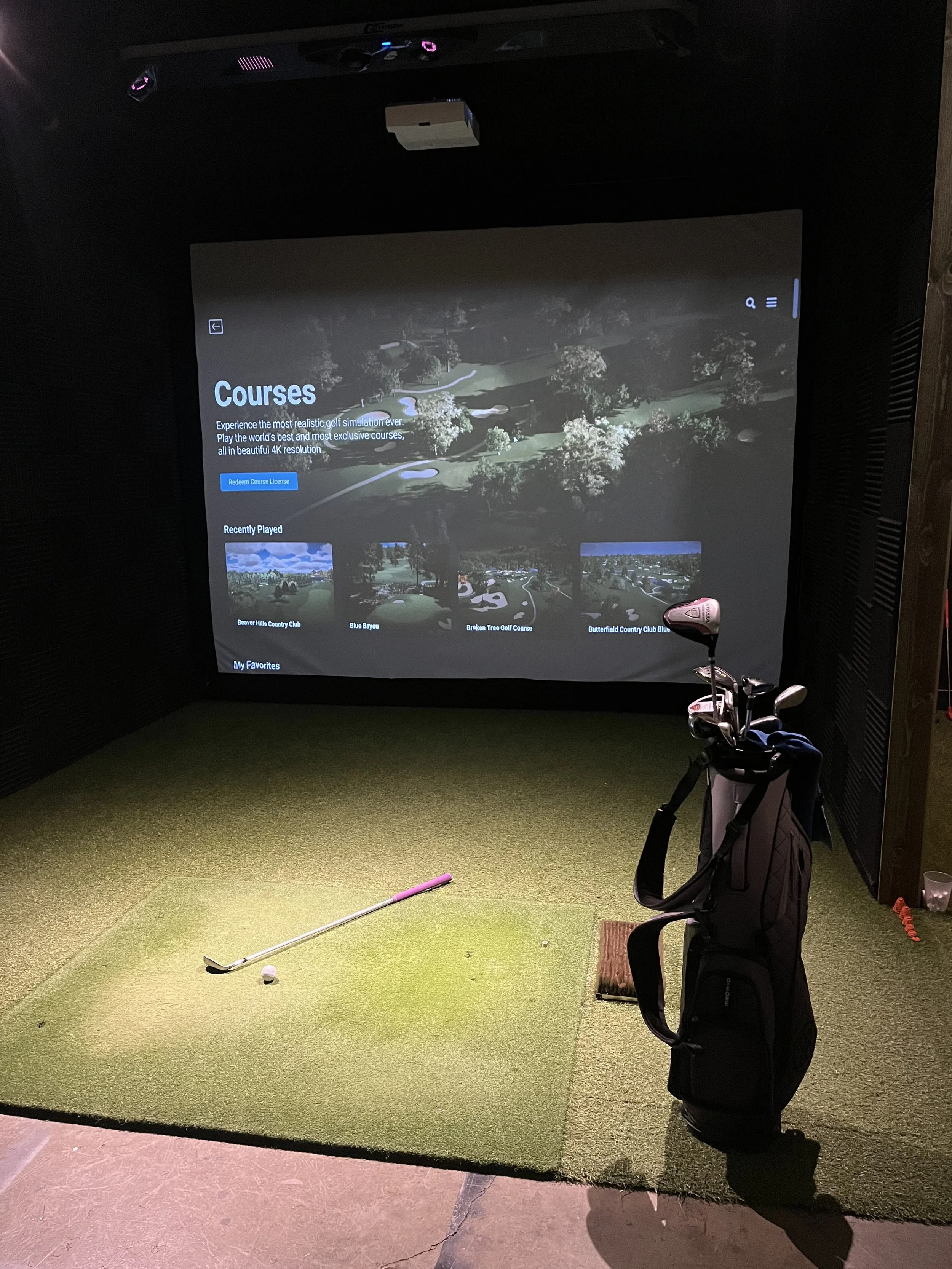 Indoor golf simulator room with a large screen displaying a golf course, golf clubs in a stand, a golf ball on the artificial turf, and a golf tee on the floor.