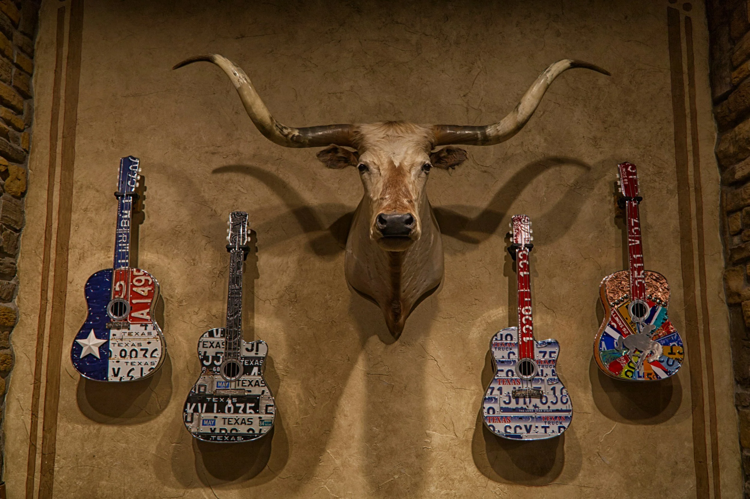 Decorative display of four guitars with license plate patterns hanging on a wall, with a mounted longhorn steer skull in the center.