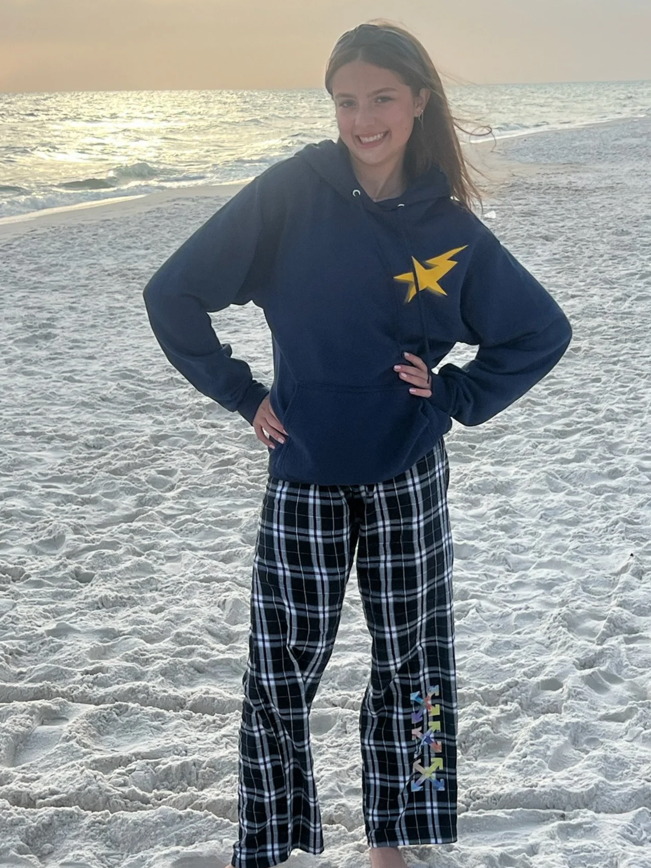 A young woman standing on a sandy beach with ocean waves in the background during sunset, smiling and posing with her hands on her hips. She is wearing a navy hoodie with a yellow star logo and black-and-white plaid pajama pants.
