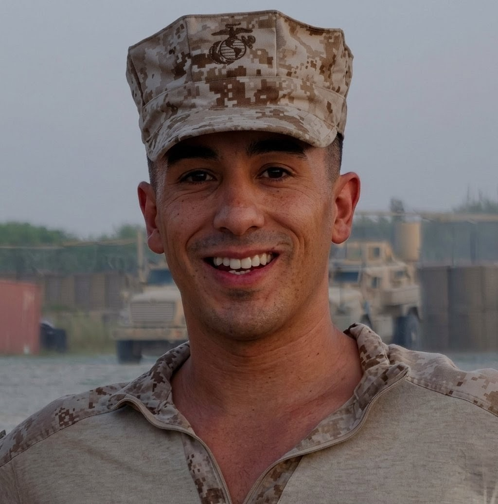 A man in a U.S. military uniform and camouflage cap smiling outdoors with a boat and water in the background.