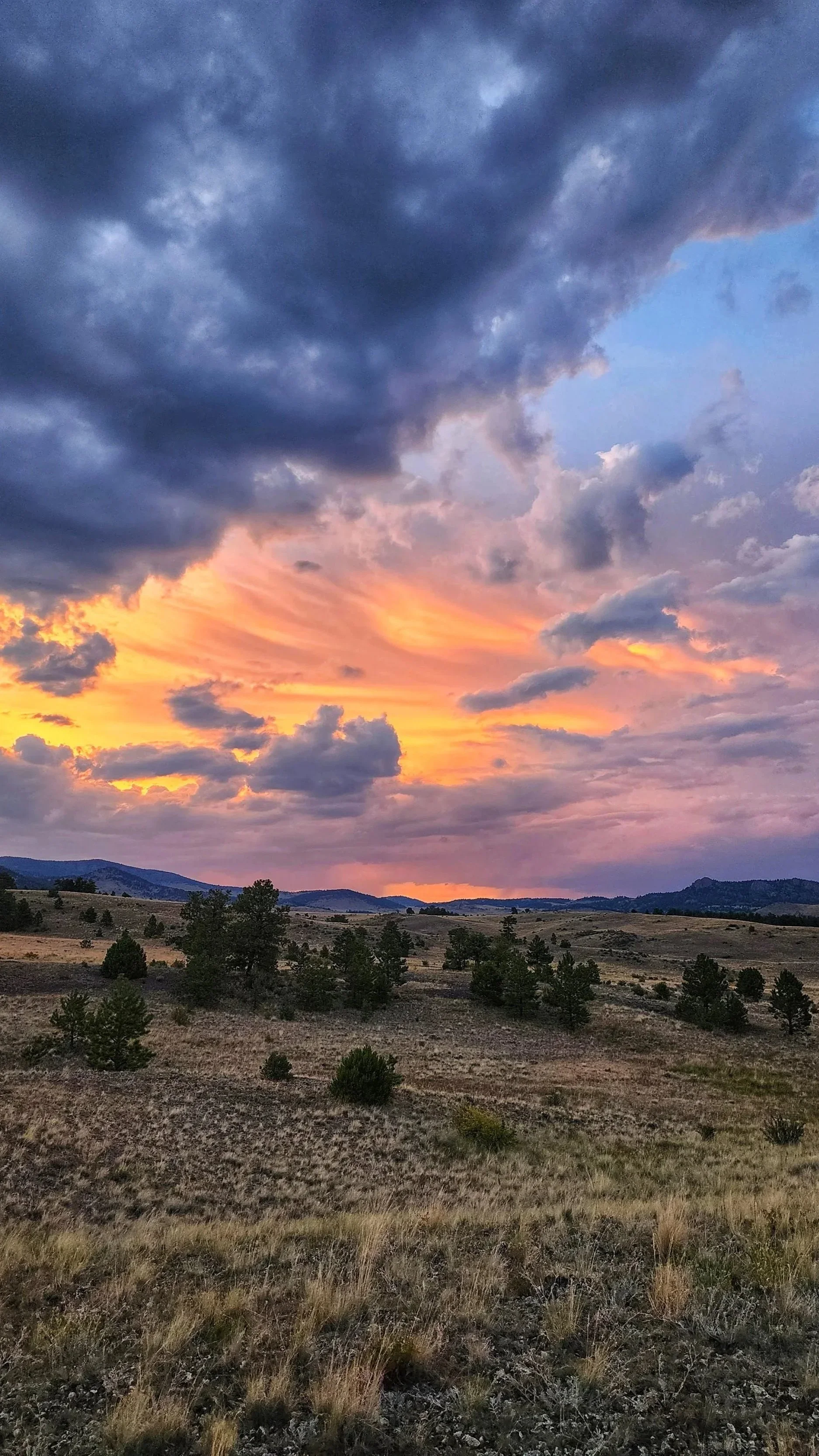 A landscape of rolling hills with sparse trees under a cloudy sky during sunset, with dark storm clouds and colorful pink, orange, and purple clouds.
