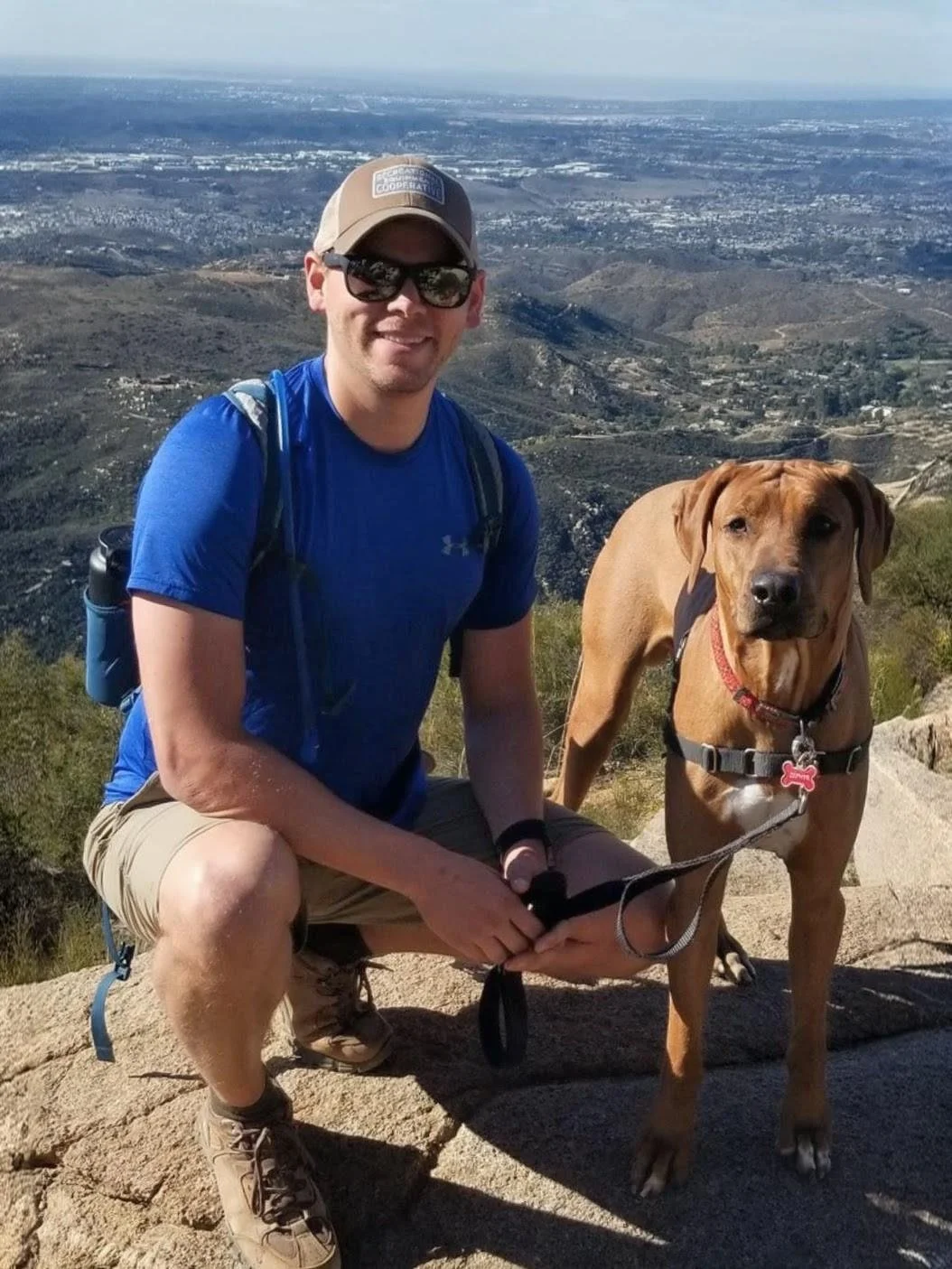 A smiling man wearing a blue shirt, shorts, hat, and sunglasses kneels next to a brown dog with a black harness on a rock ledge with a scenic mountain view in the background.