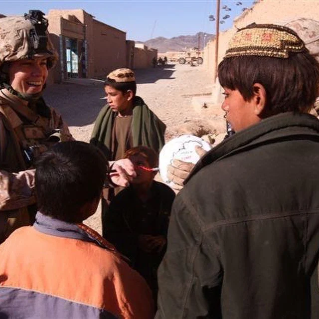Military personnel and local children in a desert village, with mountains and simple buildings in the background.