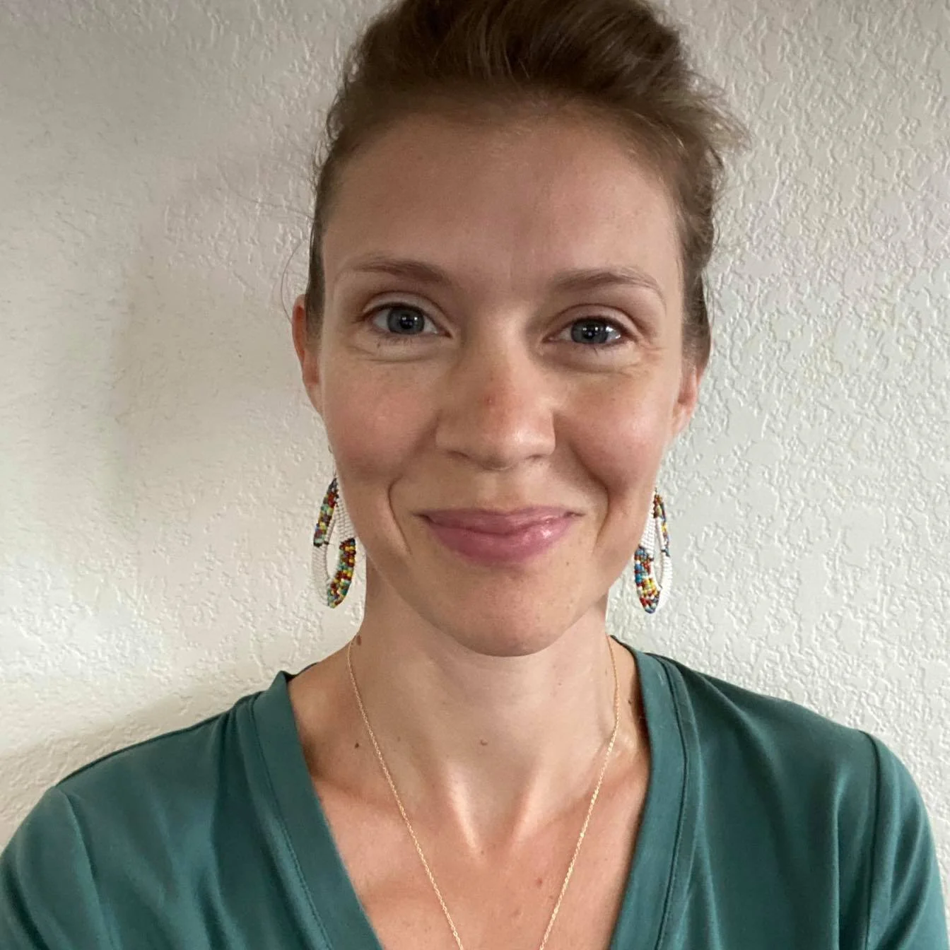 Young woman wearing beaded earrings and green shirt smiling at the camera, against a textured wall background.