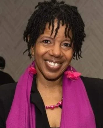 Middle aged woman smiling with short dreadlocks wearing a pink scarf and earrings.