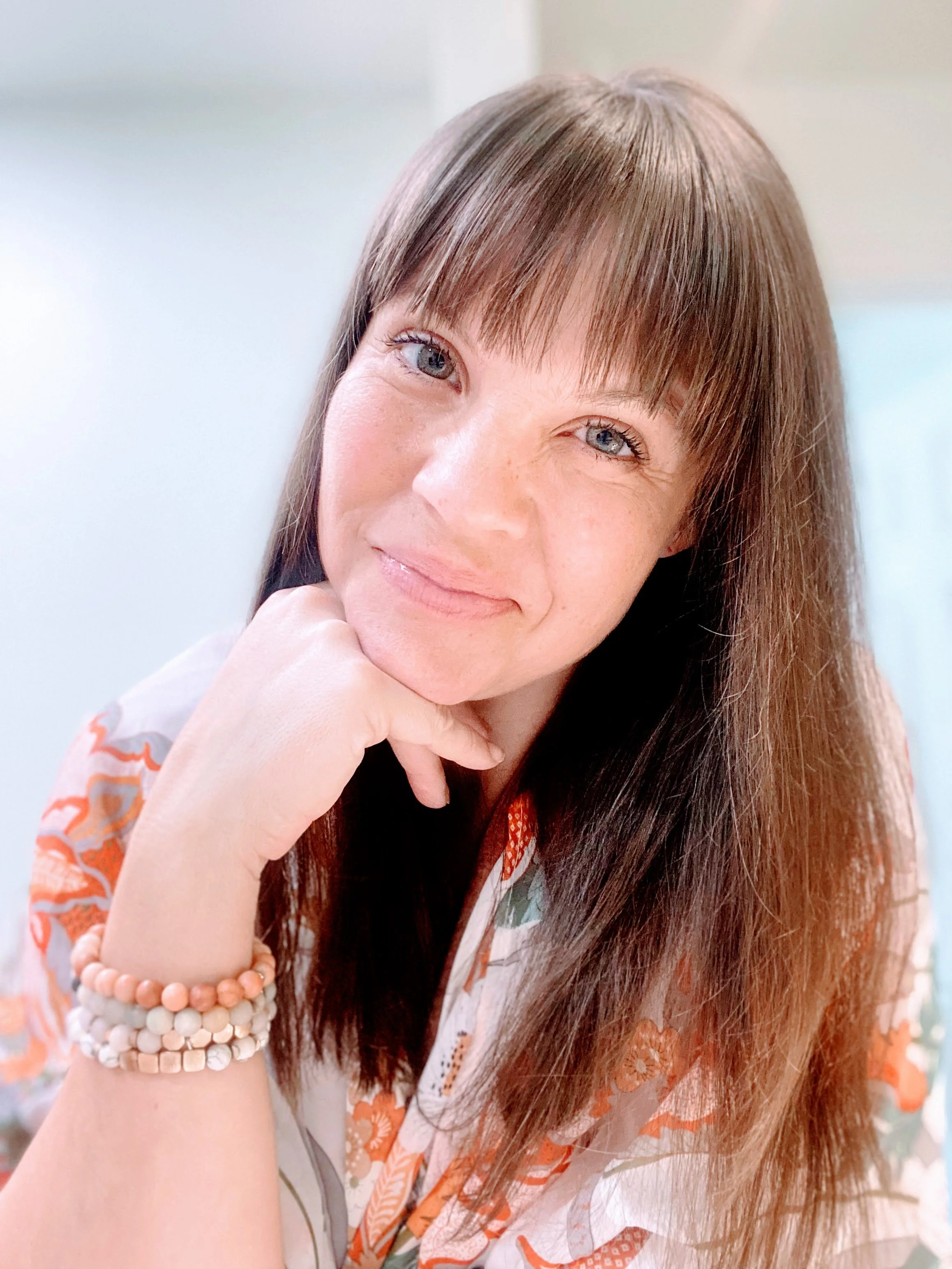 A woman with long brown hair and bangs, wearing a patterned top, smiles at the camera. She has her hand under her chin and is wearing beaded bracelets.