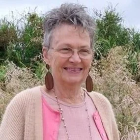 Elderly woman smiling outdoors, wearing a pink shirt, beige sweater, large brown earrings, and glasses, with greenery in the background.