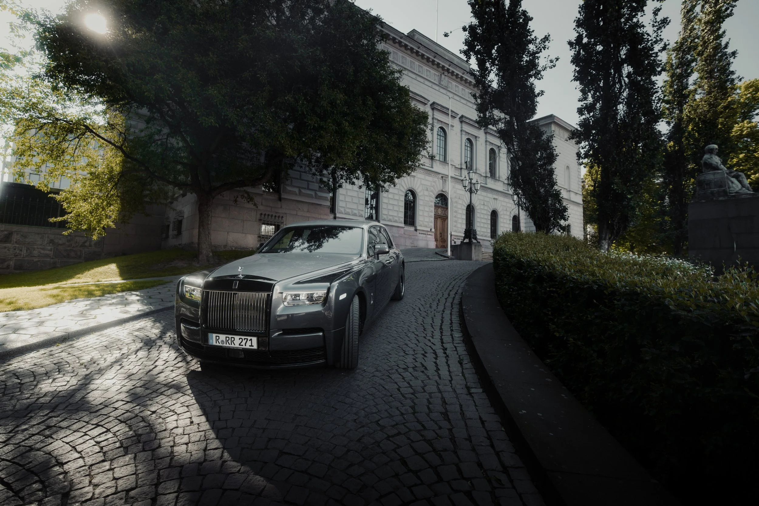 A black luxury car parked on a cobblestone street in front of an elegant white building with large windows and ornate architectural details, surrounded by trees and statues.