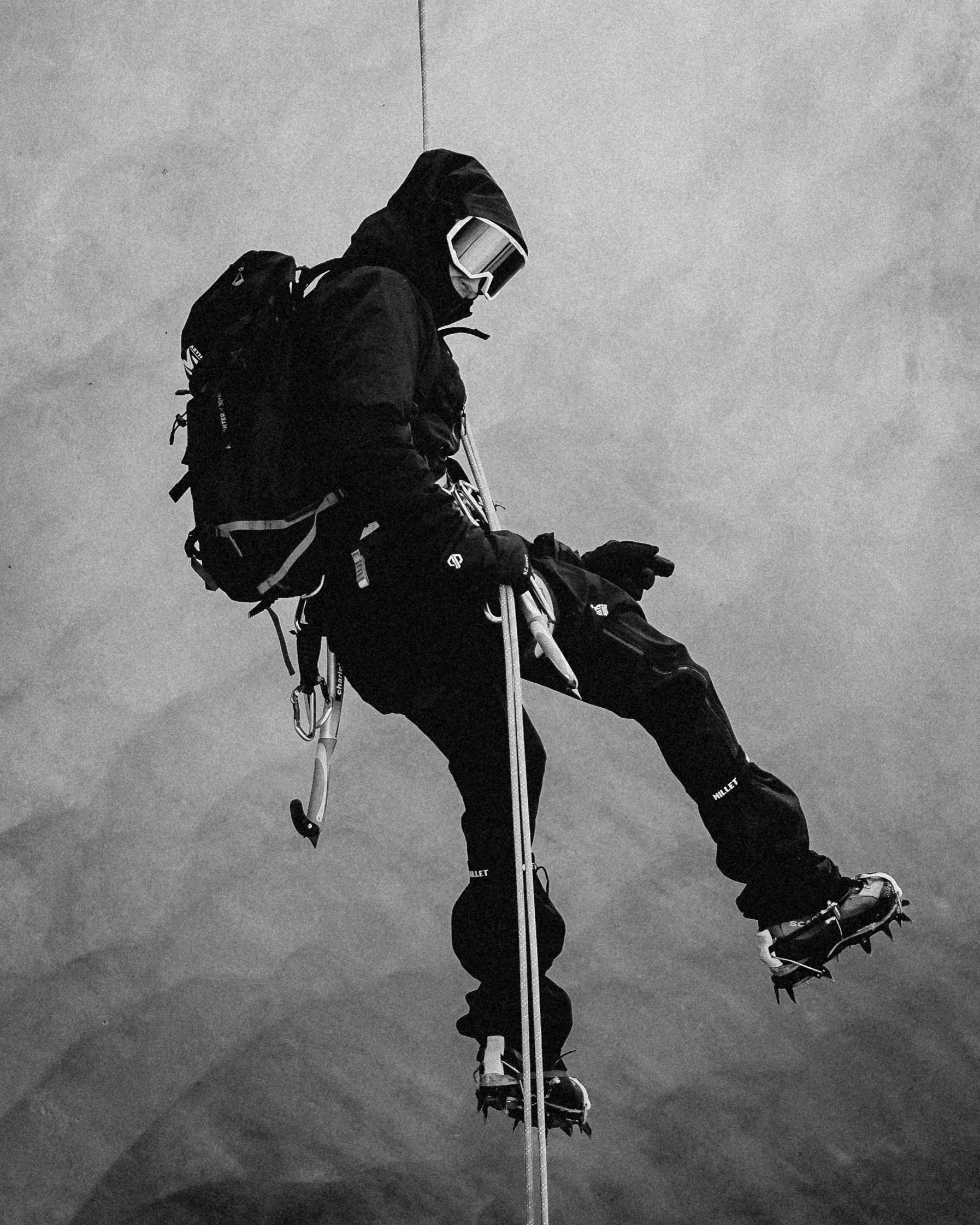 A person dressed in black mountaineering gear, including goggles, a hood, backpack, and crampons on their boots, is climbing a steep icy surface.