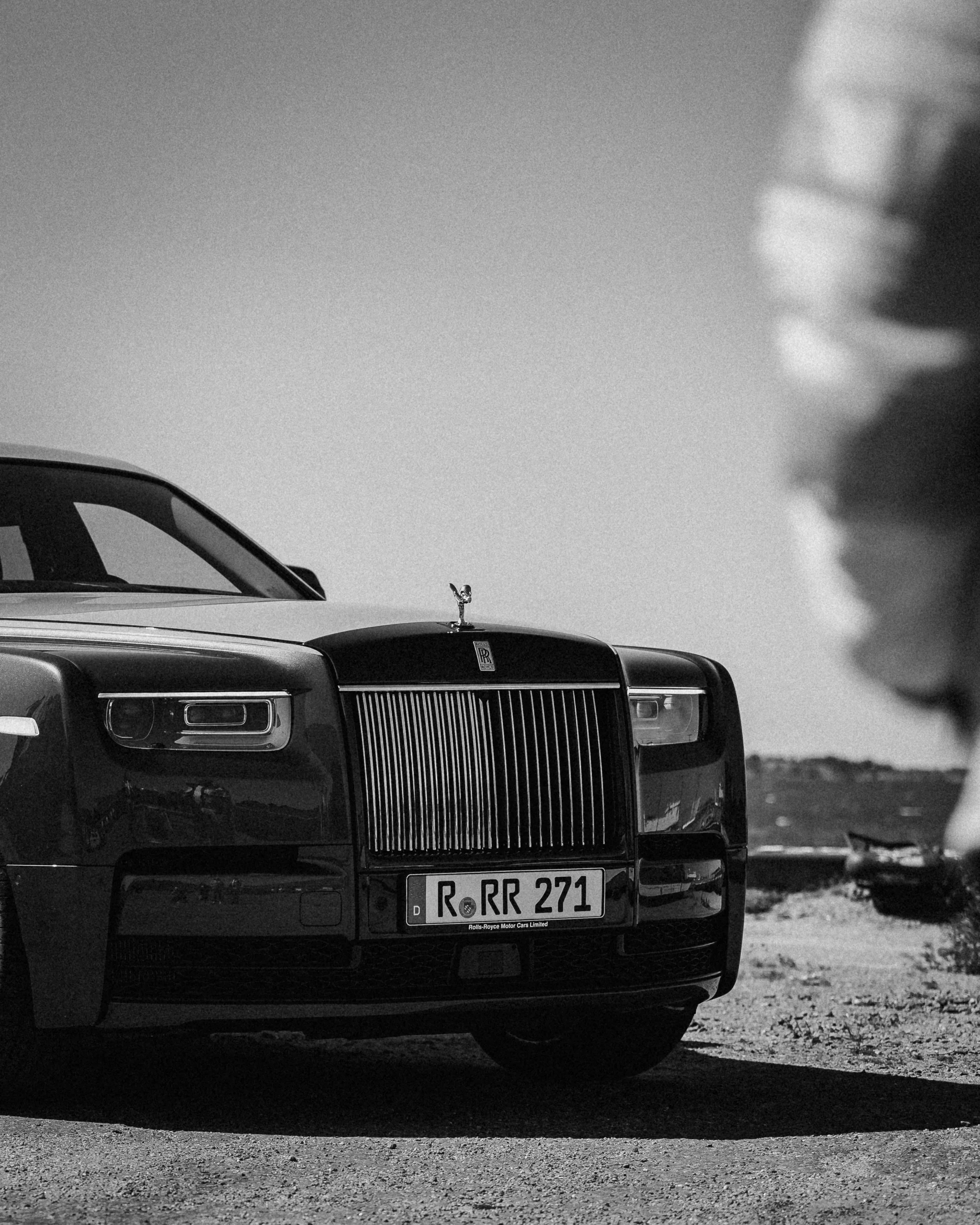 Black Rolls-Royce car with a front grille and hood ornament visible, parked outdoors on a dirt surface with a blurred background, black and white photo.