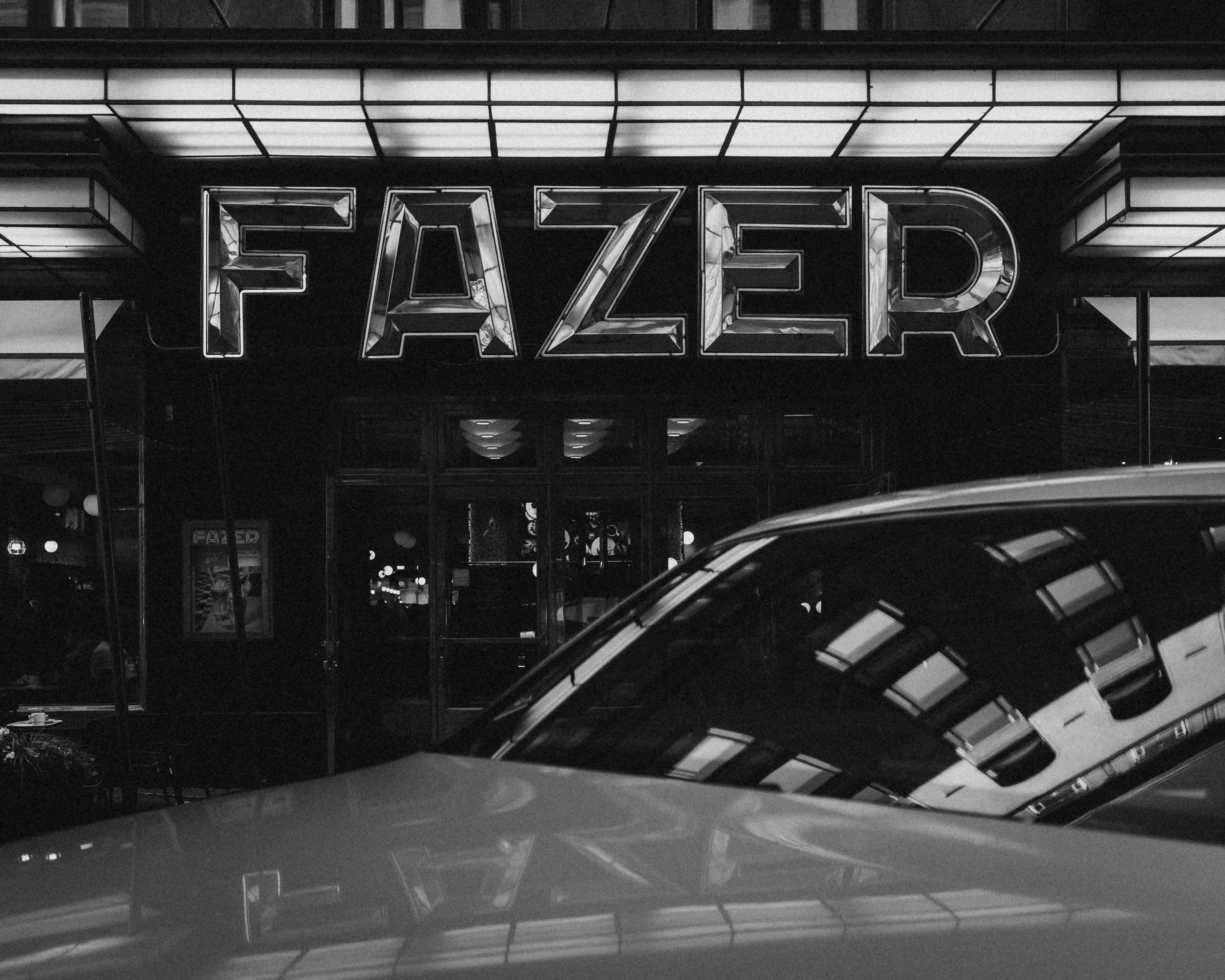 The exterior of a fuzzy, illuminated sign reading 'FAZER' in large capital letters outside a building, with reflections of nearby structures visible.