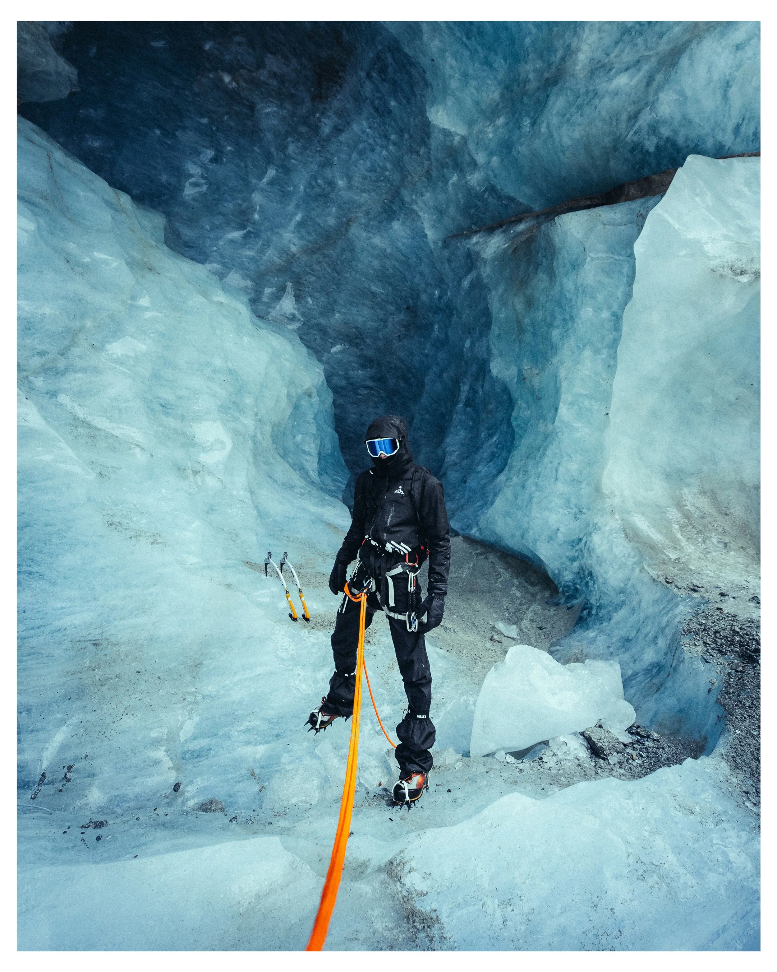 A person dressed in black mountaineering gear with ice axes and crampons on icy terrain inside a glacier cavern.