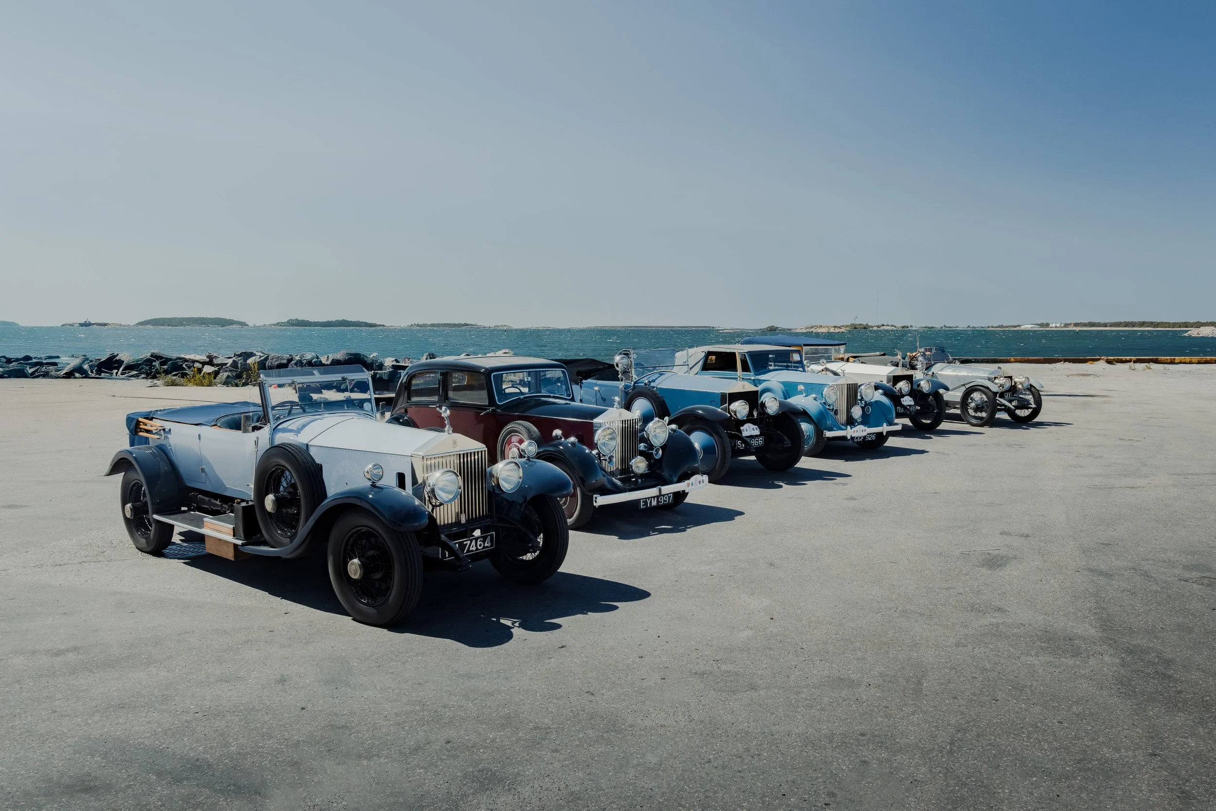 A line of six vintage cars parked on a concrete surface near the water with a rocky breakwater and blue sky in the background.