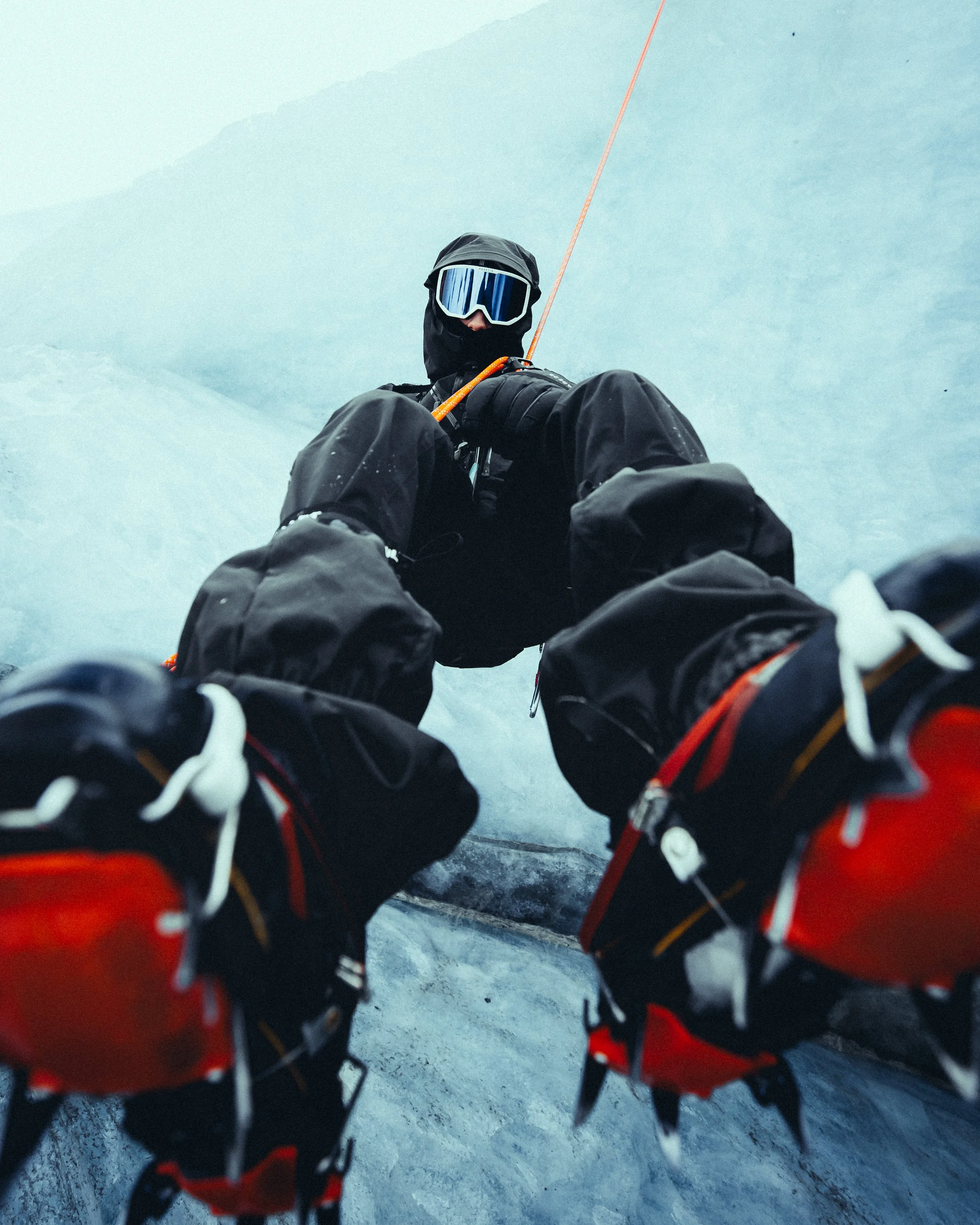 A climber dressed in black climbing gear using an ice axe on a snowy mountain slope, wearing goggles, with snow-covered mountains in the background.