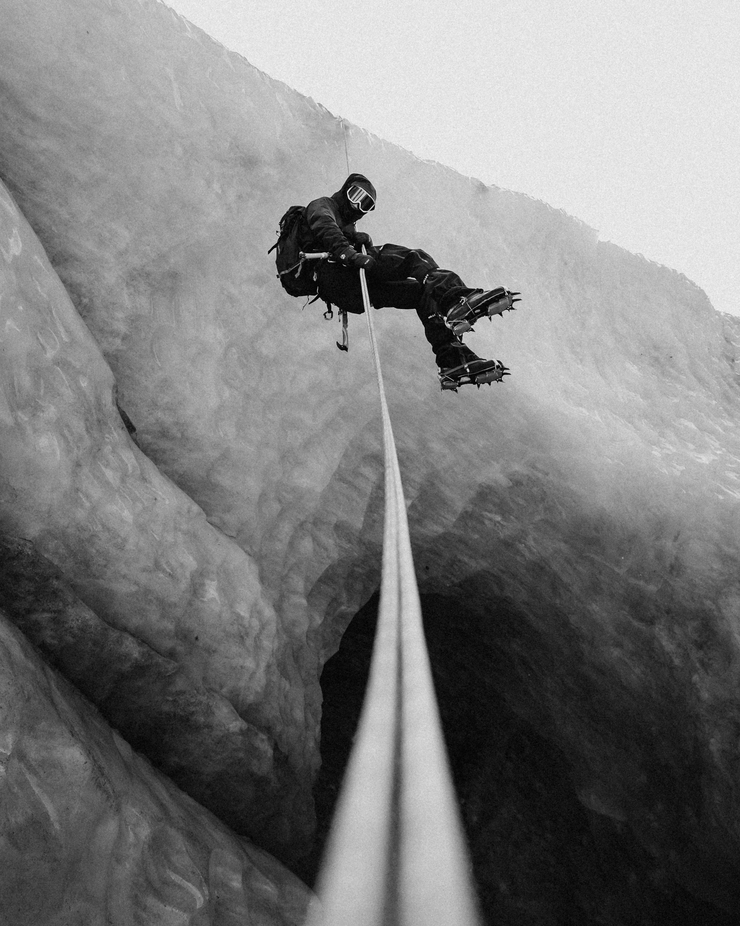A black and white photo of a climber sitting on a rope spanning a deep crevice in a mountain, wearing protective gear.