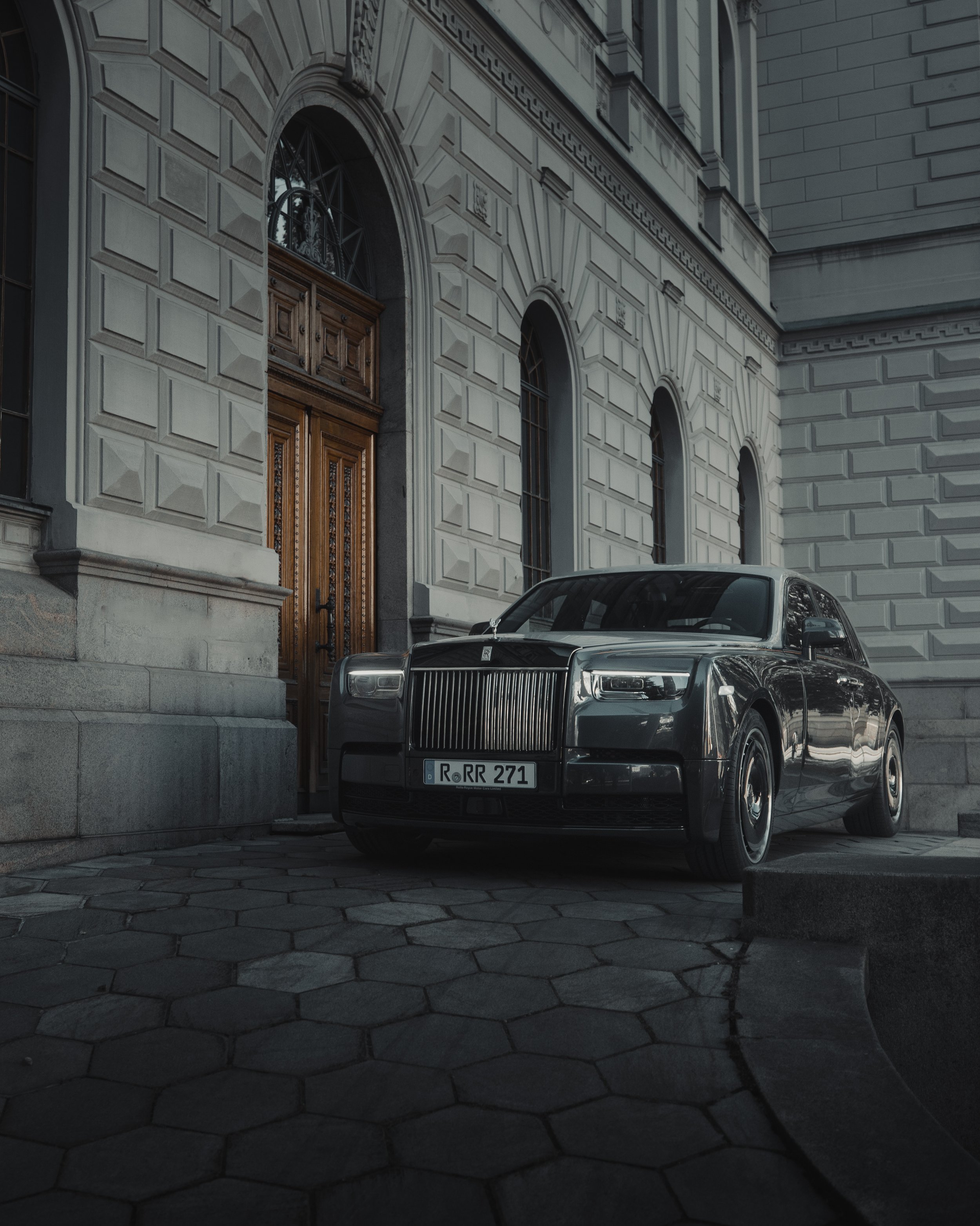 A black luxury Rolls-Royce car parked in front of a historic stone building with arched windows and a wooden door.