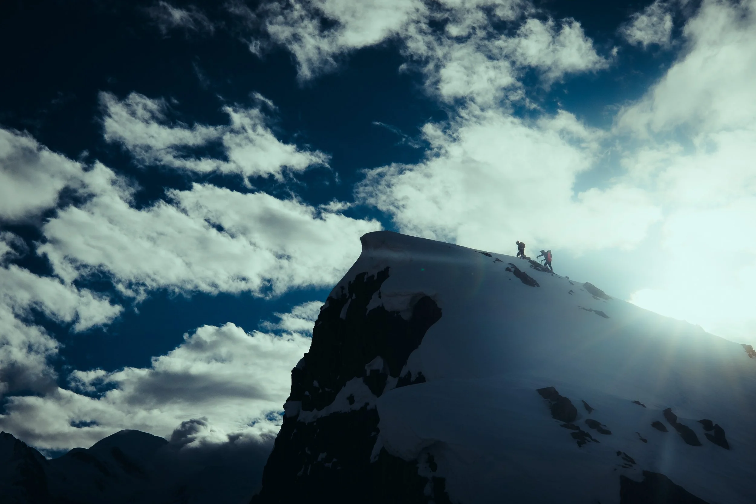 Two climbers ascending a snow-covered mountain peak in silhouette against a partly cloudy sky with the sun shining.