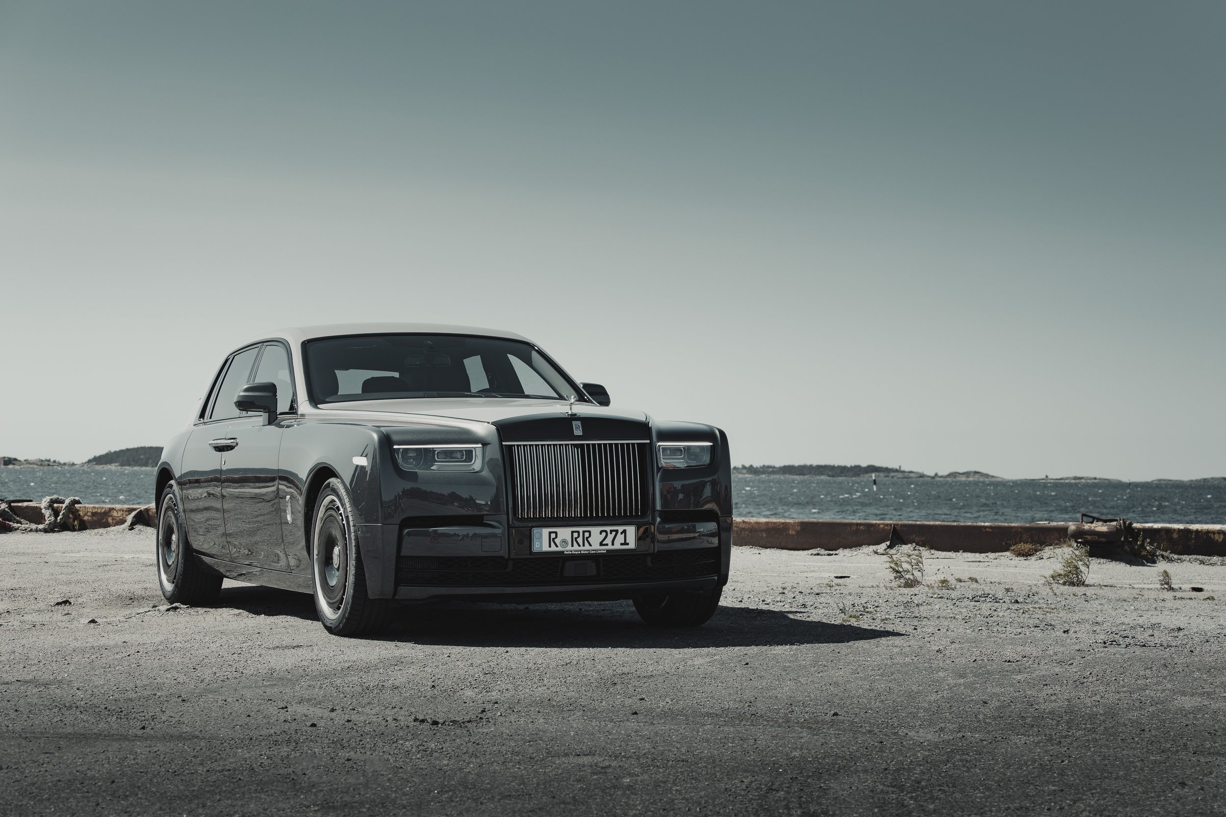 A black luxury Rolls-Royce car parked near the water on a paved area with a cloudy sky and distant islands in the background.