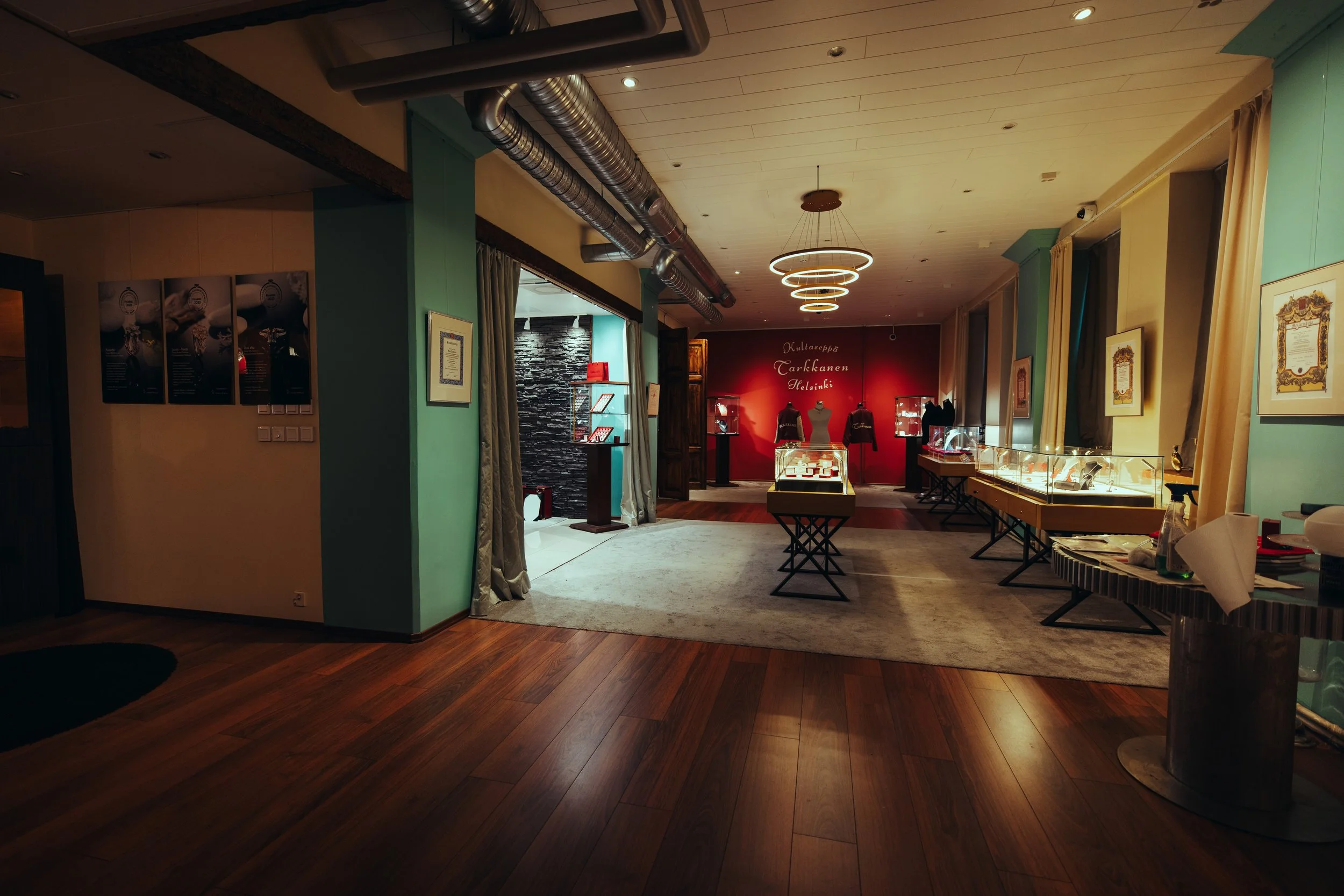 An exhibition room displaying jewelry and accessories in glass cases, a red accented wall with mannequins, and artwork on the walls, suggesting a museum or gallery setting.