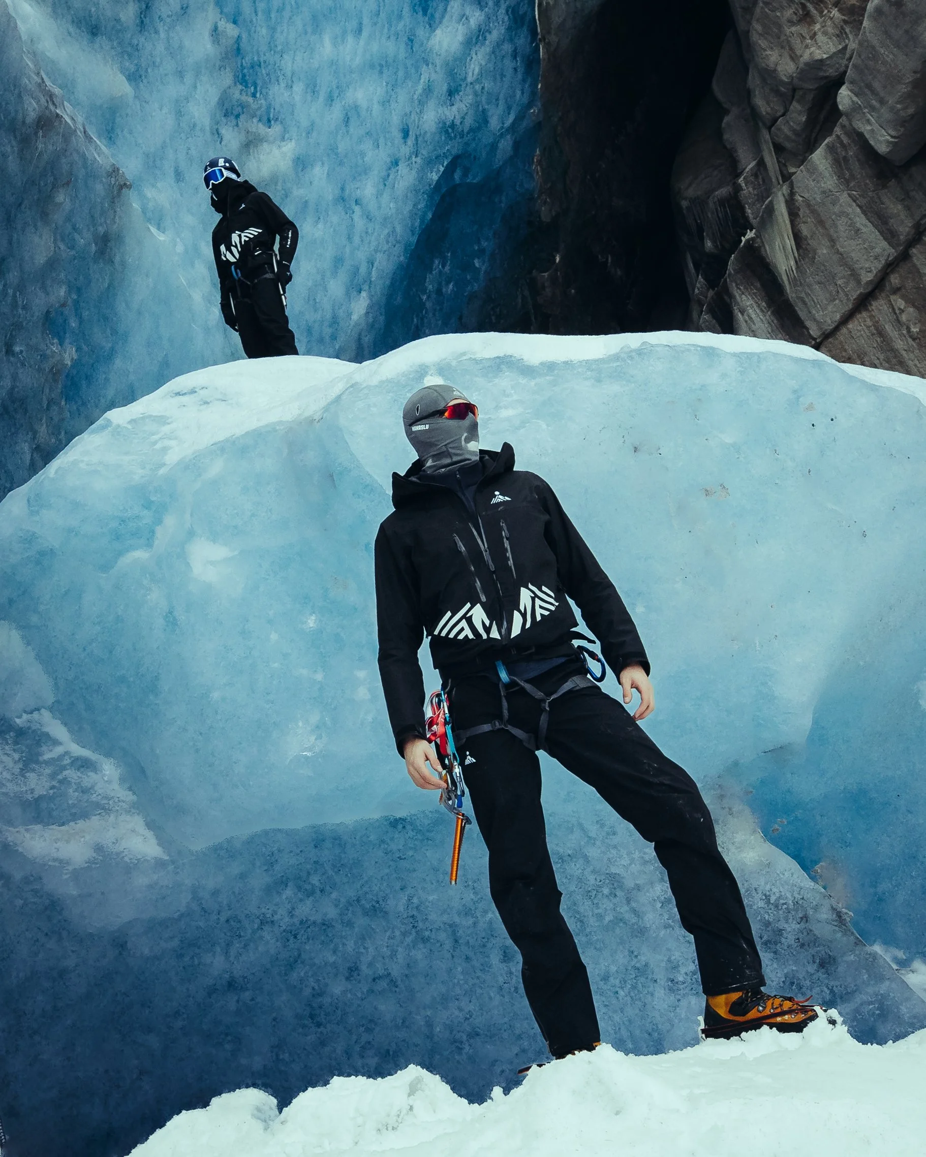 Two climbers in black ice climbing gear on a glacier with blue ice and rocky cliffs in the background.