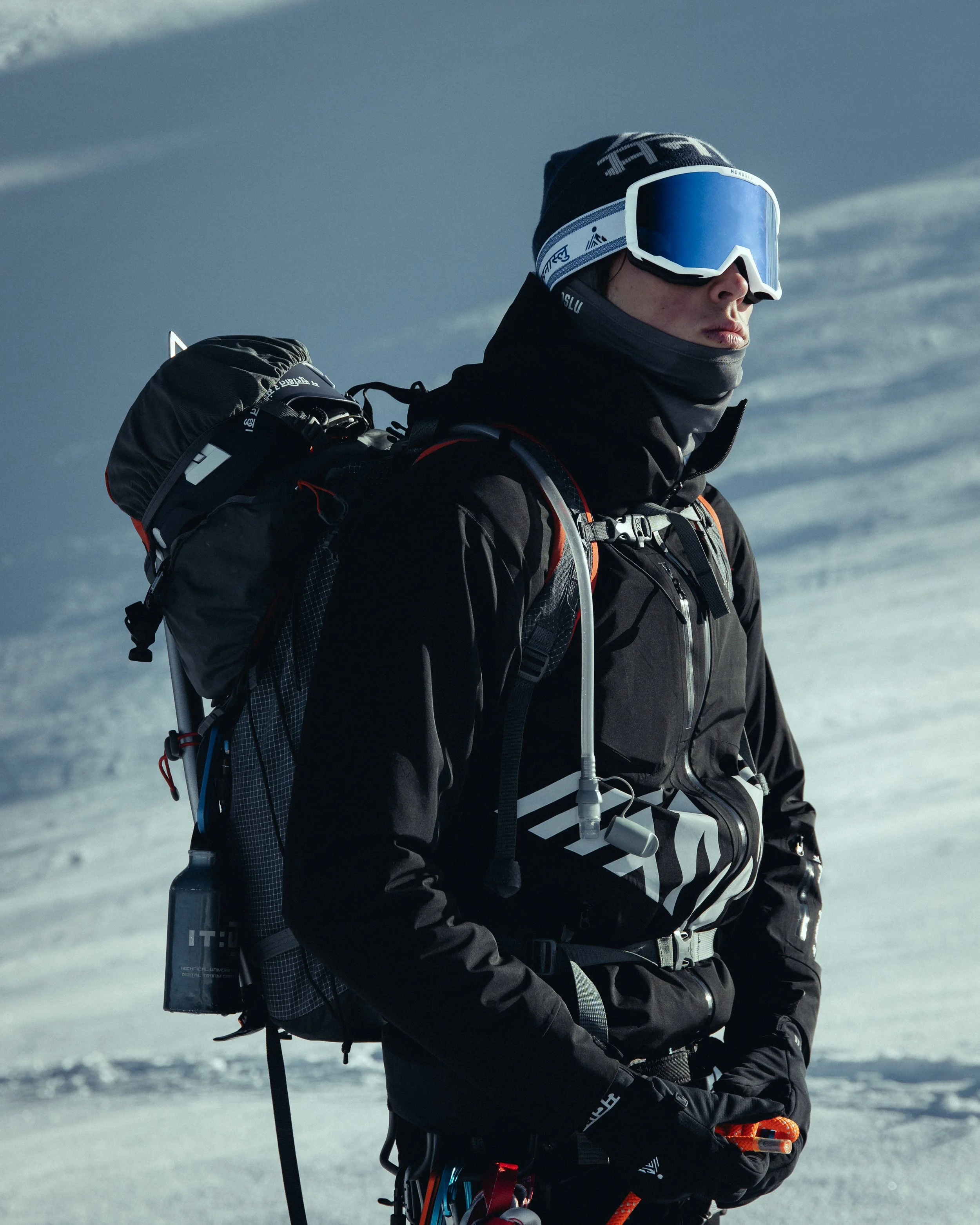A man in winter gear, wearing a black jacket, black gloves, blue ski goggles, and a black beanie, standing on snow-covered terrain with a backpack equipped with tools and gear.