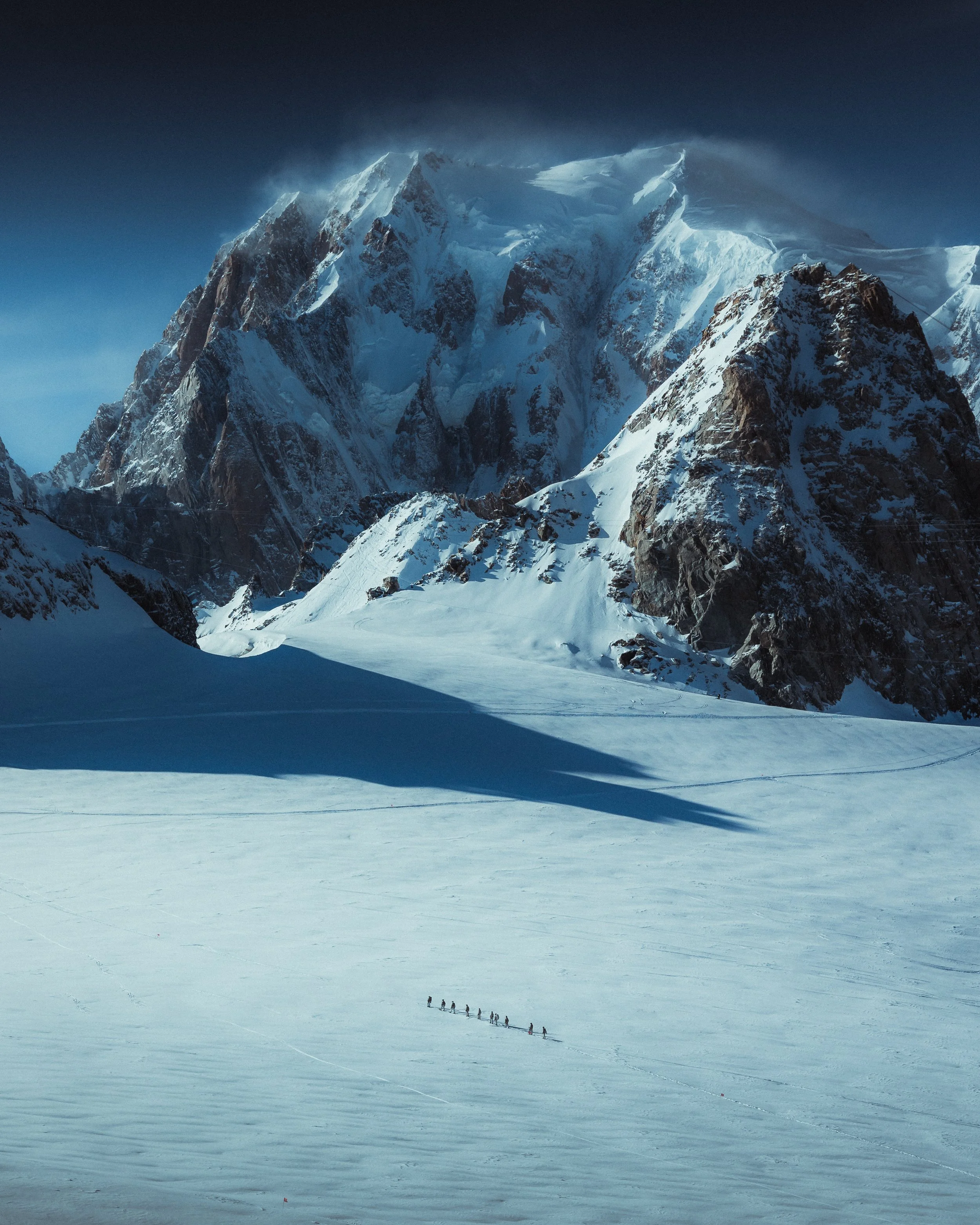 A snow-covered mountain range with a group of people hiking across the snowfield below.