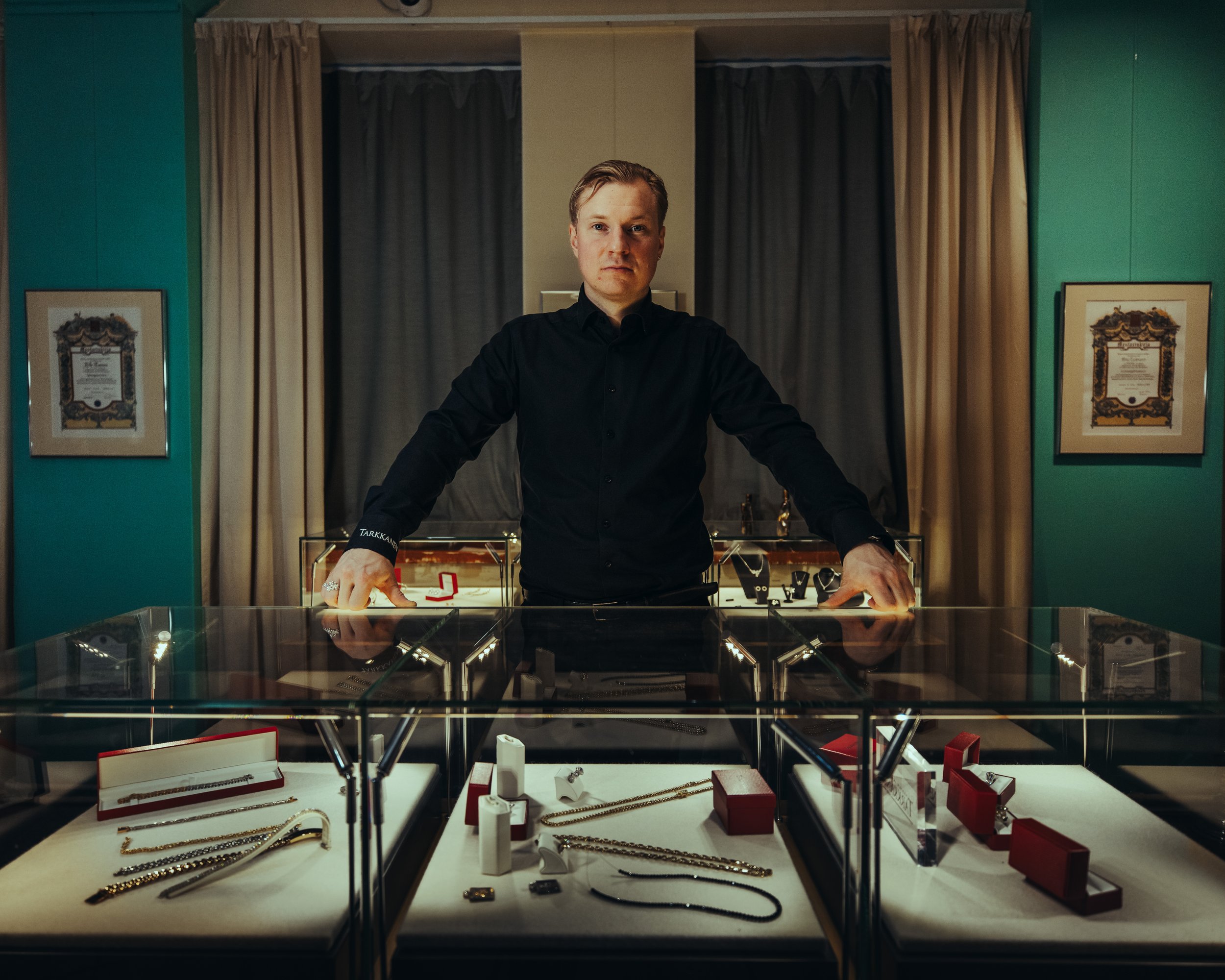 A man is standing behind a jewelry display case with various necklaces, rings, and watches inside, in a store with framed certificates on the teal walls and curtains in the background.