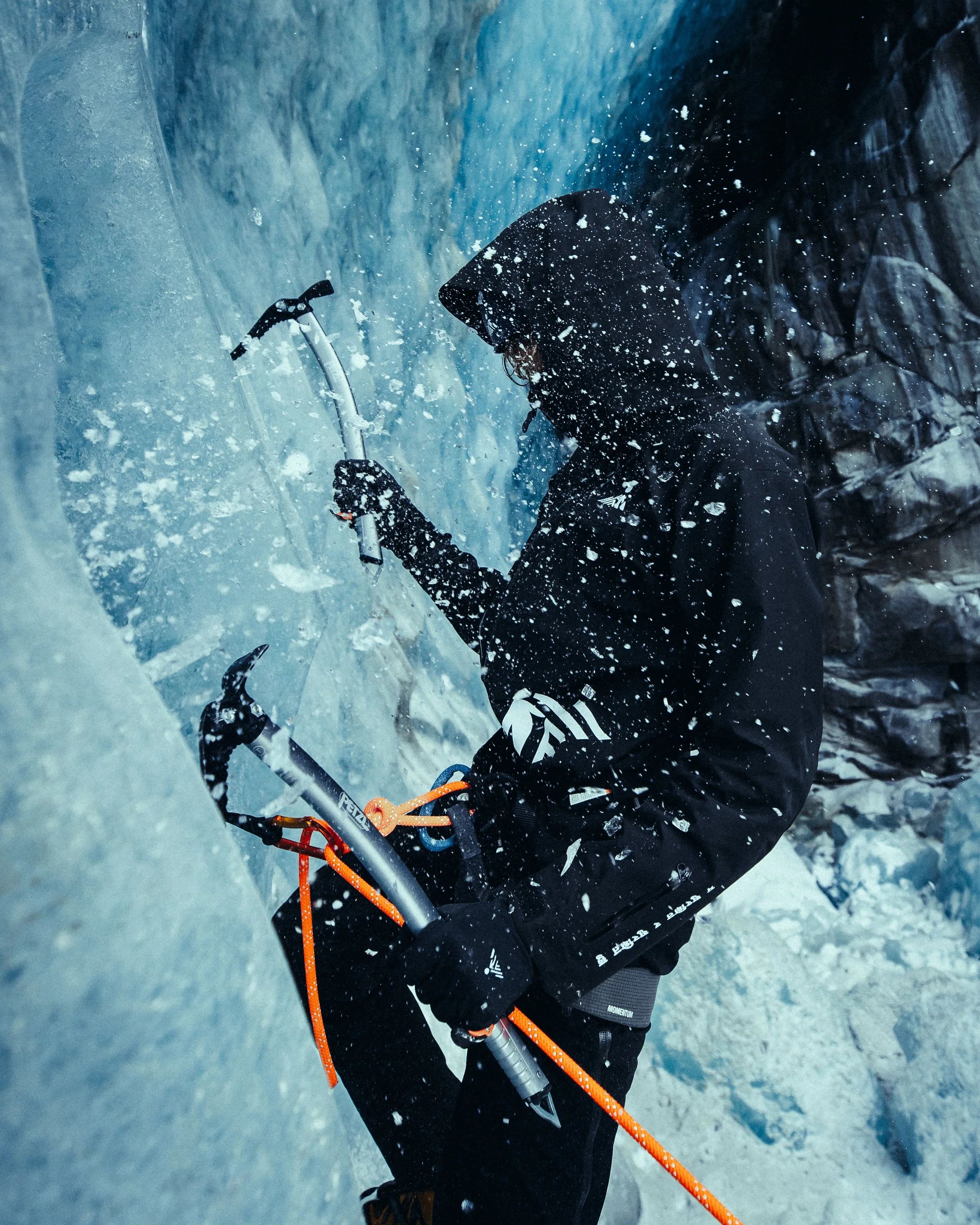 A person dressed in black winter clothing climbing an icy surface with ice axes, surrounded by snow and ice splashes.