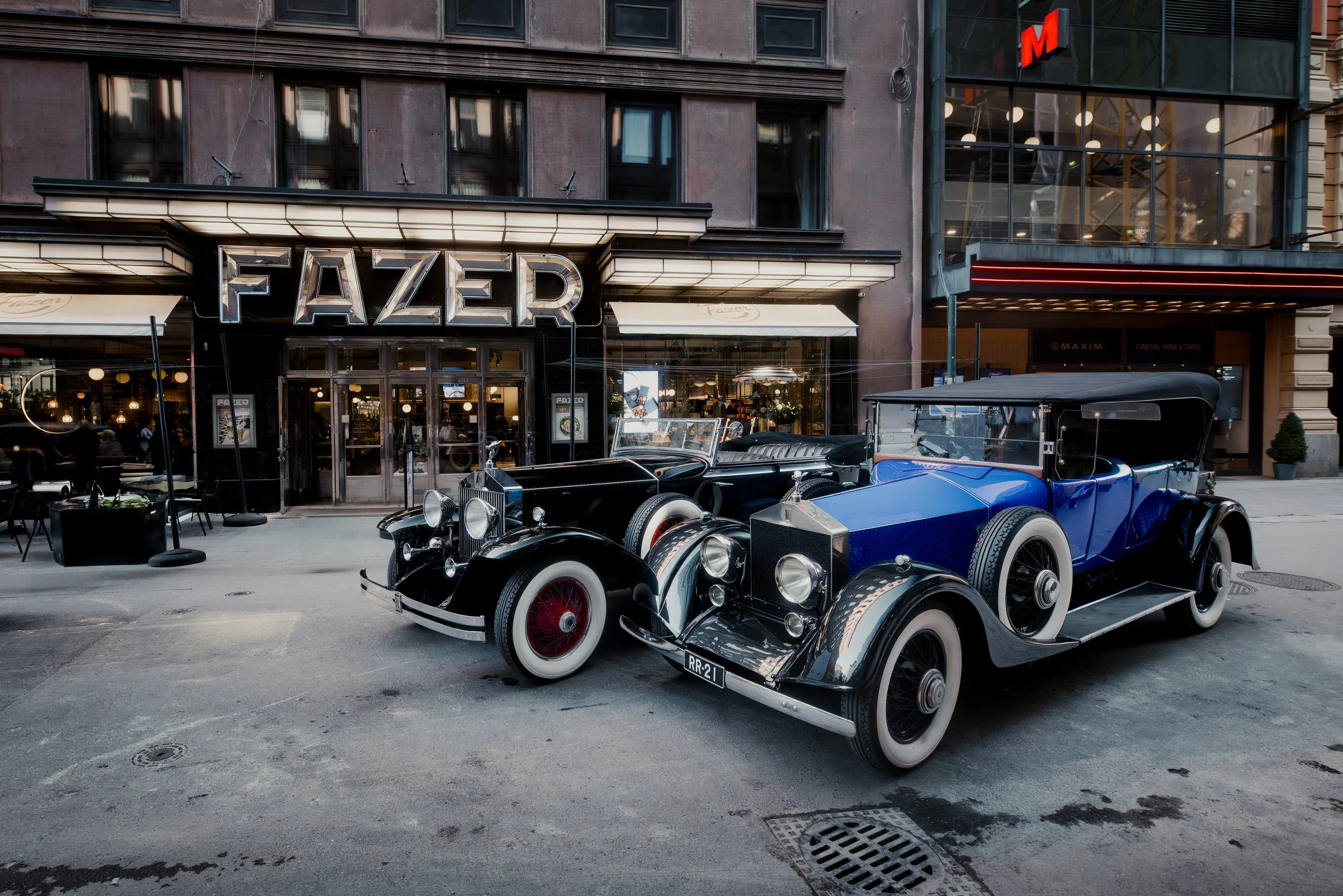 Two vintage cars parked in front of a restaurant called Fazer on a city street.