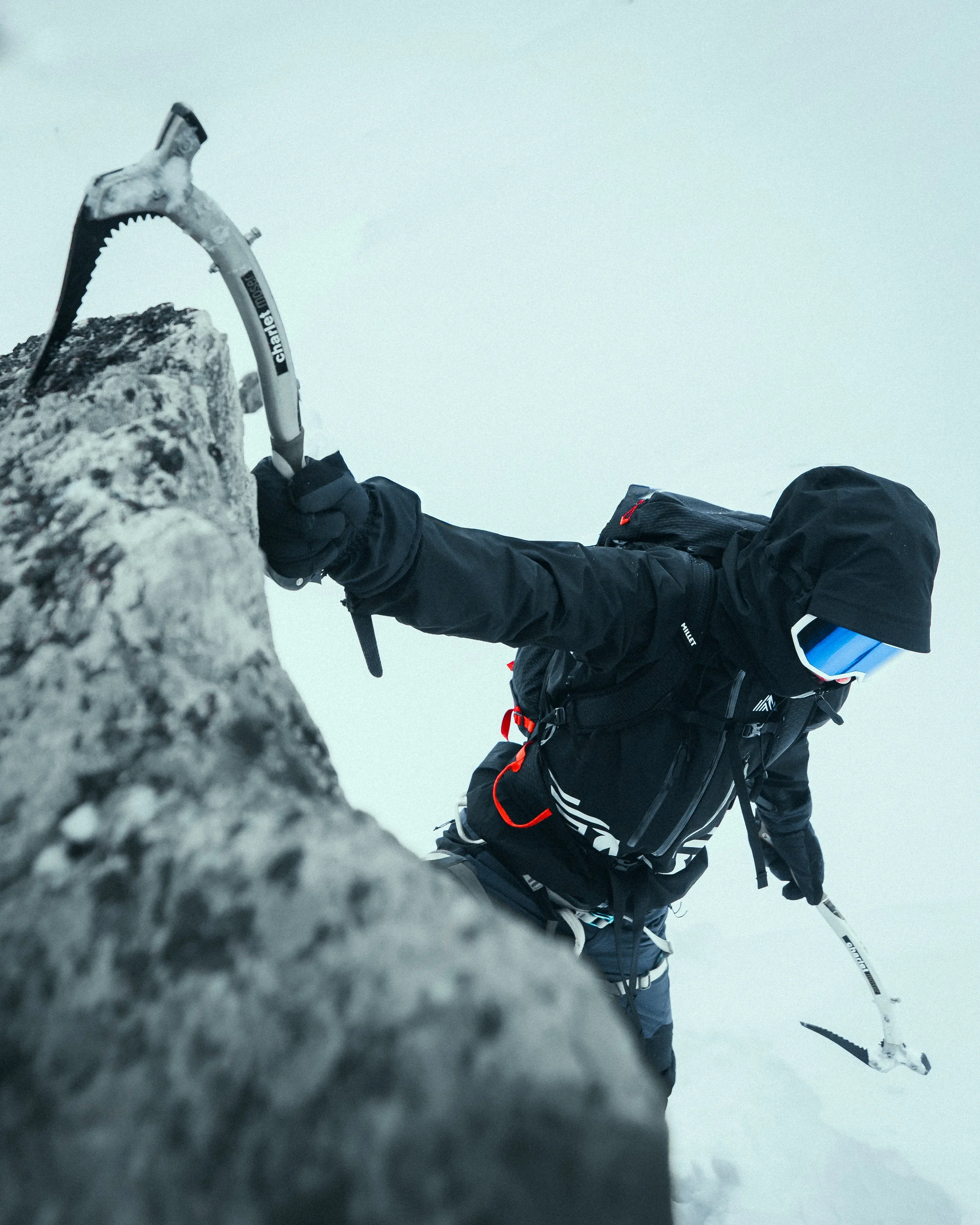 Mountain climber in black winter gear ascending icy rock face with ice axe, foggy weather, and snow.