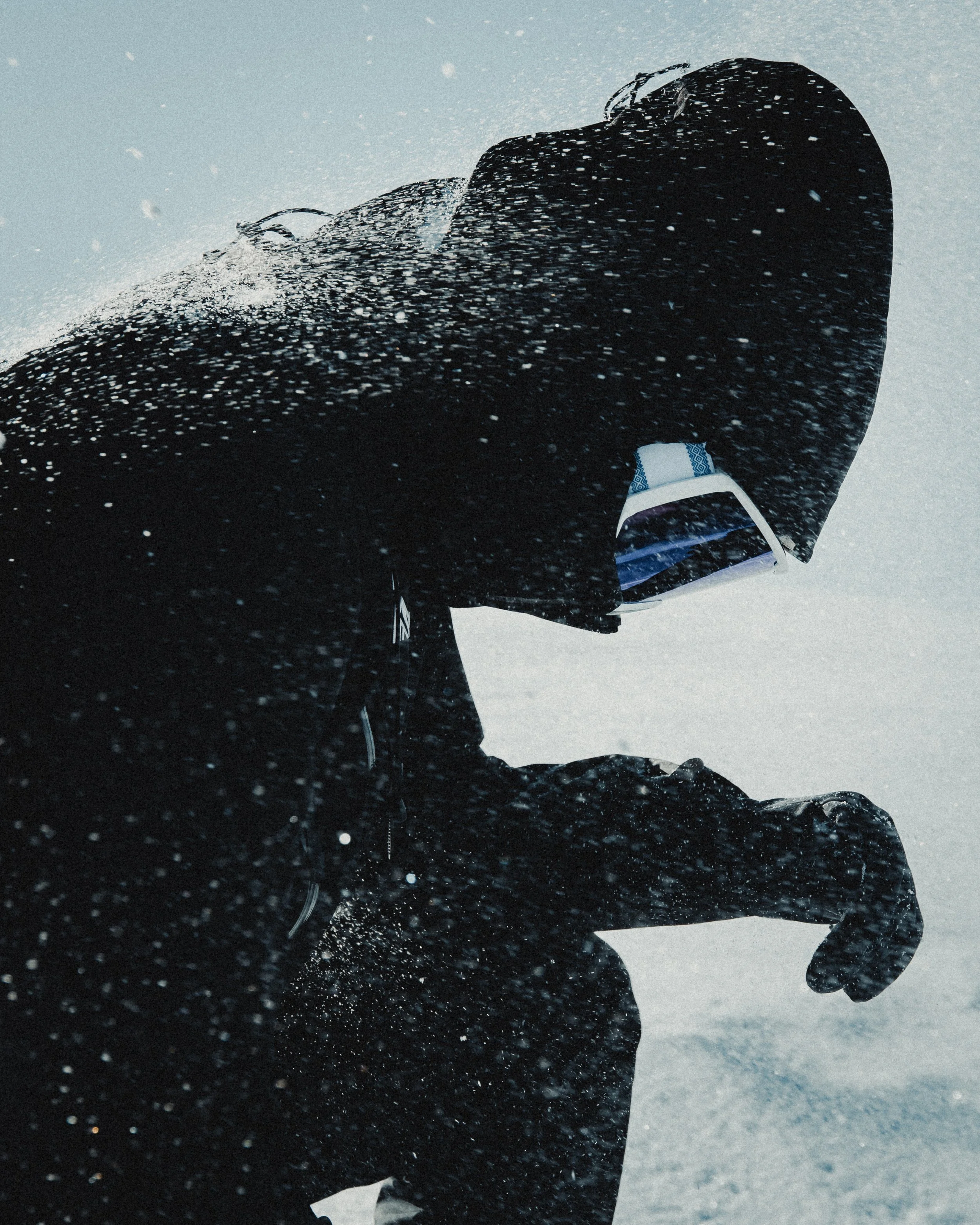 Person dressed in black winter jacket with hood and glasses crouching in snow during a snowstorm.