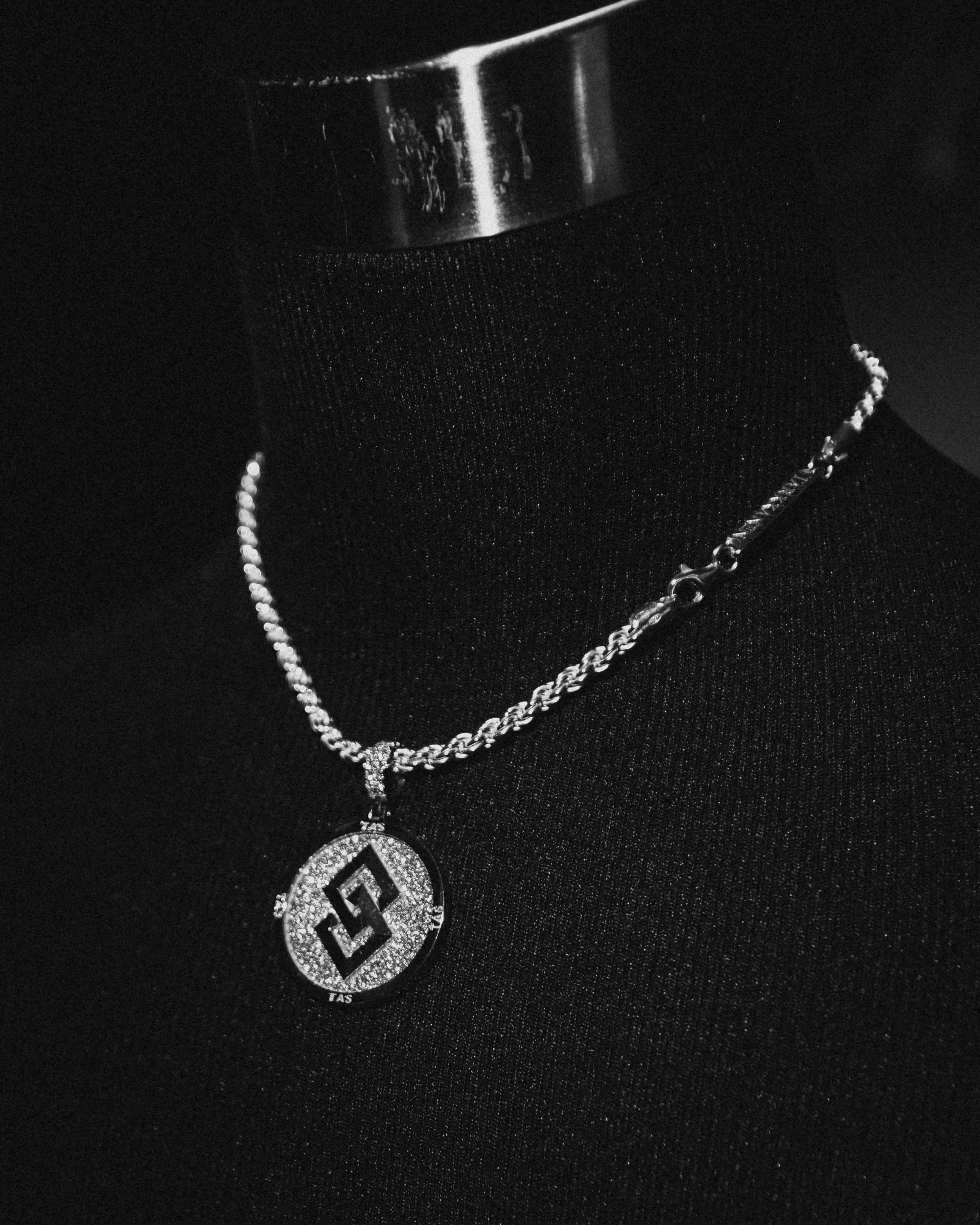 A silver chain necklace with a circular pendant, displayed on a black textured mannequin or background.