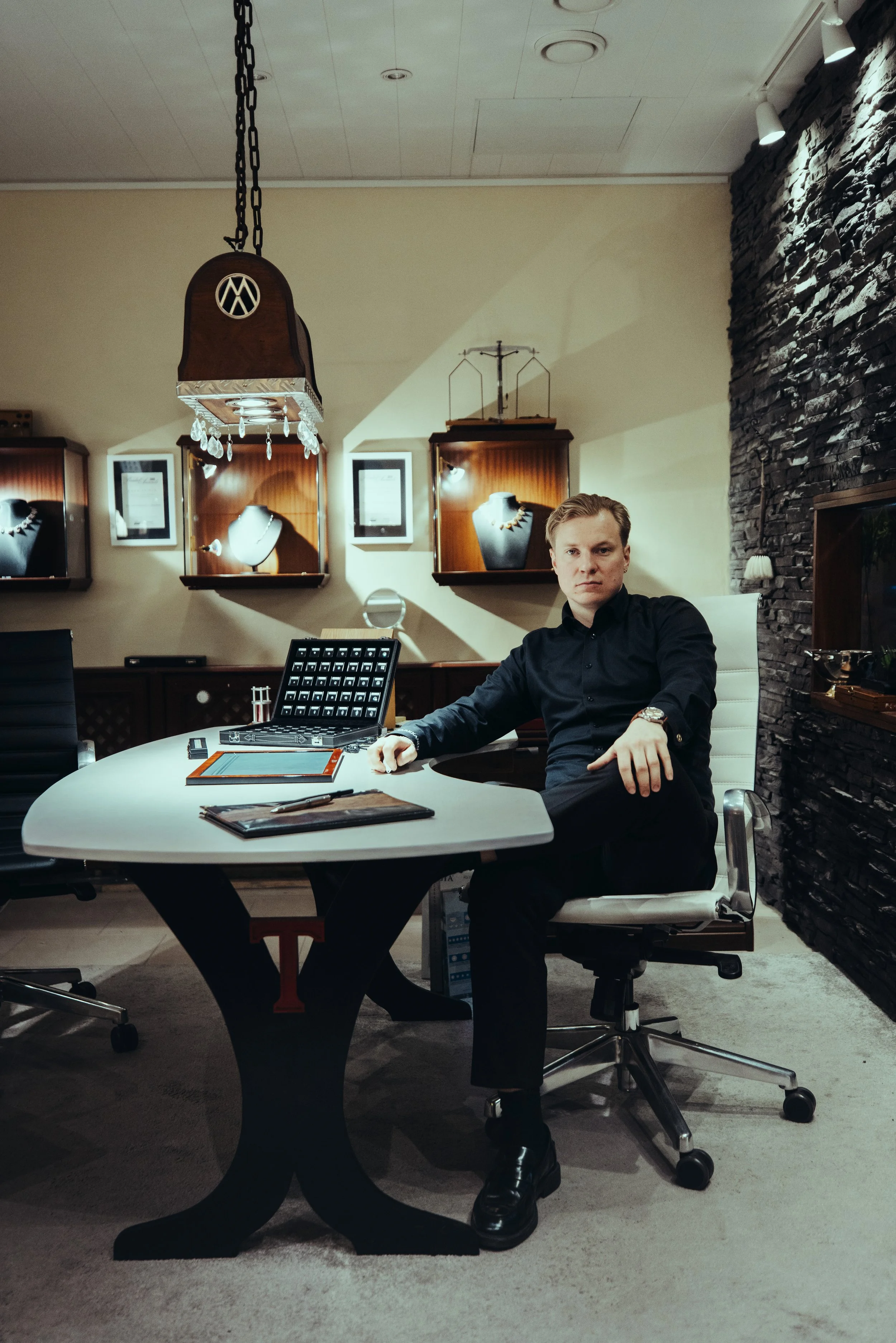 A man sitting at a modern white office desk in an elegant room with jewelry display cases and dark stone wall.