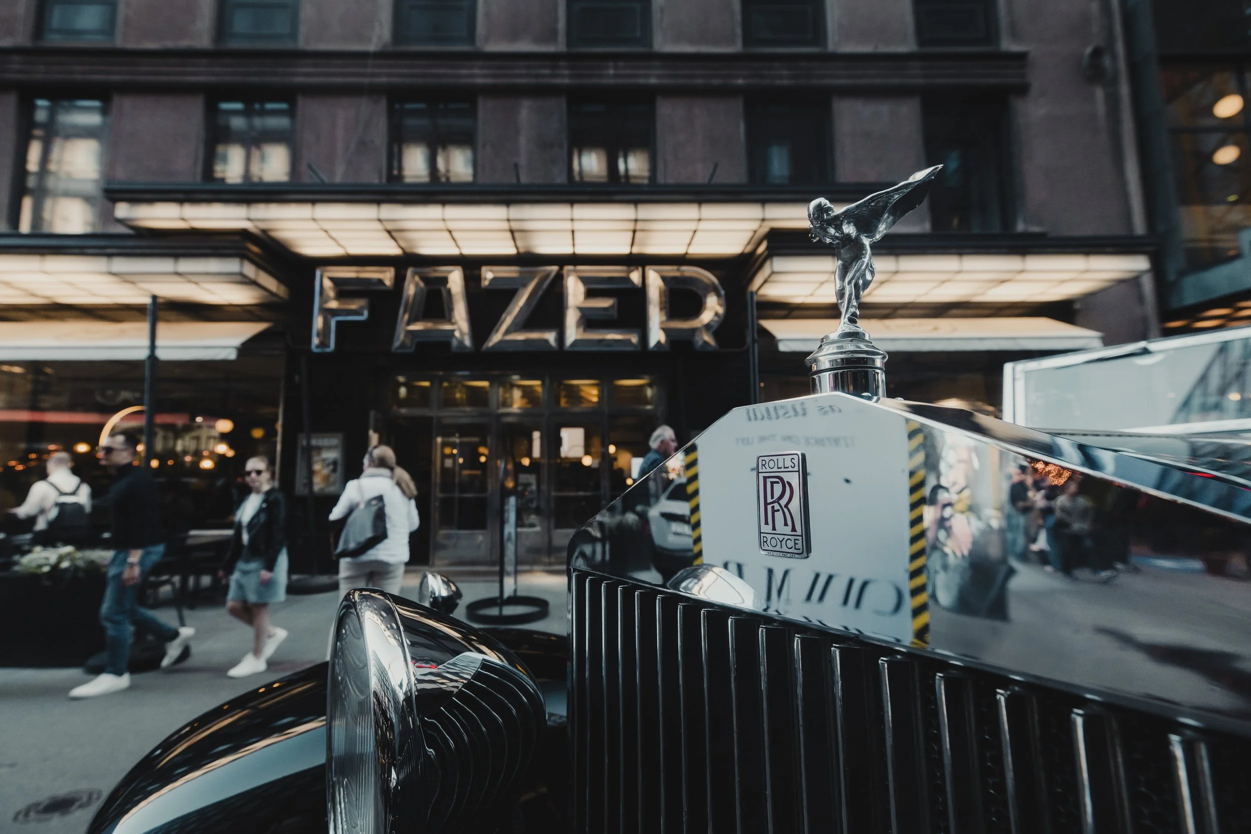 Close-up of a silver Rolls Royce with the Spirit of Ecstasy hood ornament, parked in front of the FAZER building with people walking on the sidewalk.