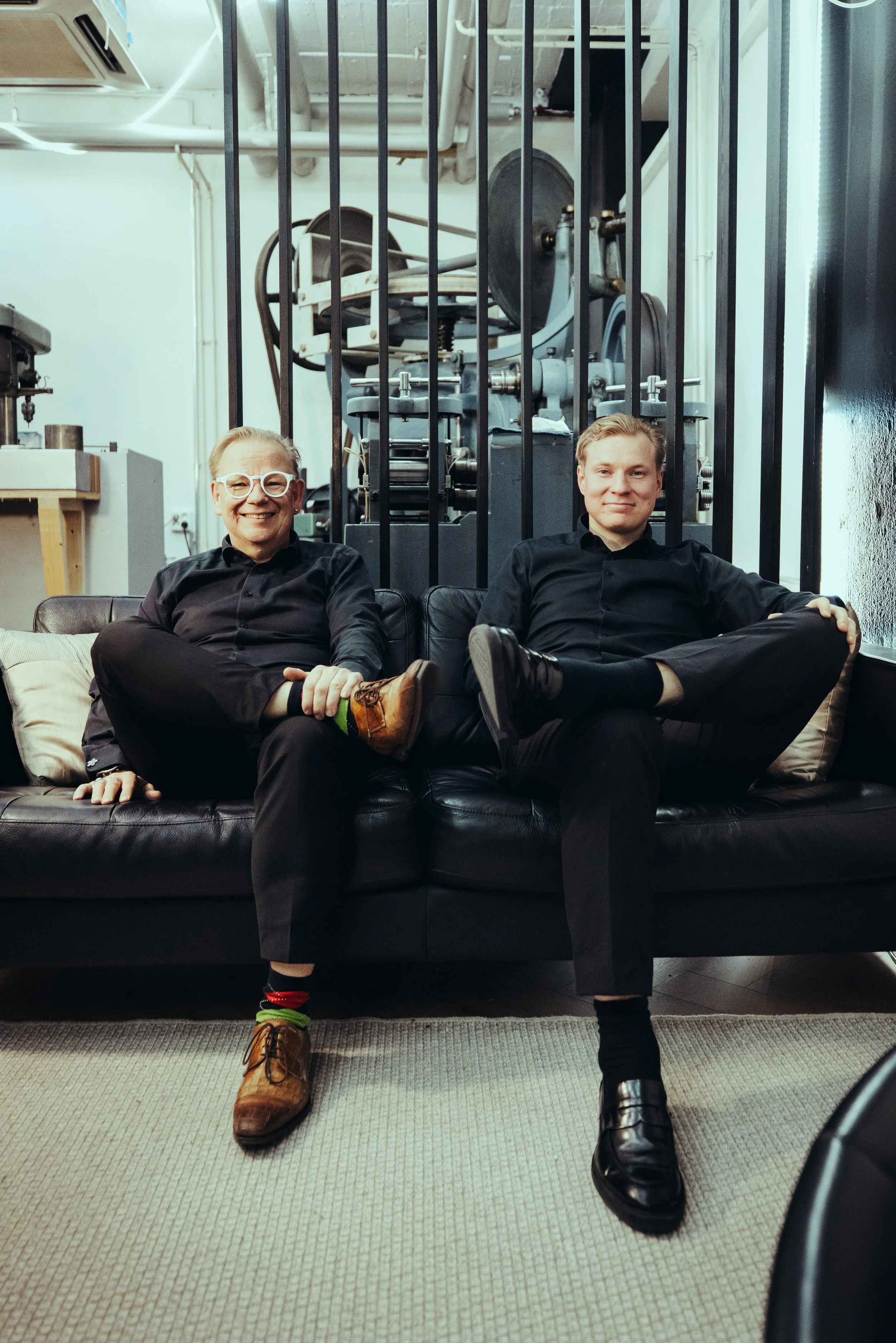Two men sitting on a black leather couch in a modern industrial-style office, smiling, with large machinery visible through a glass partition behind them.