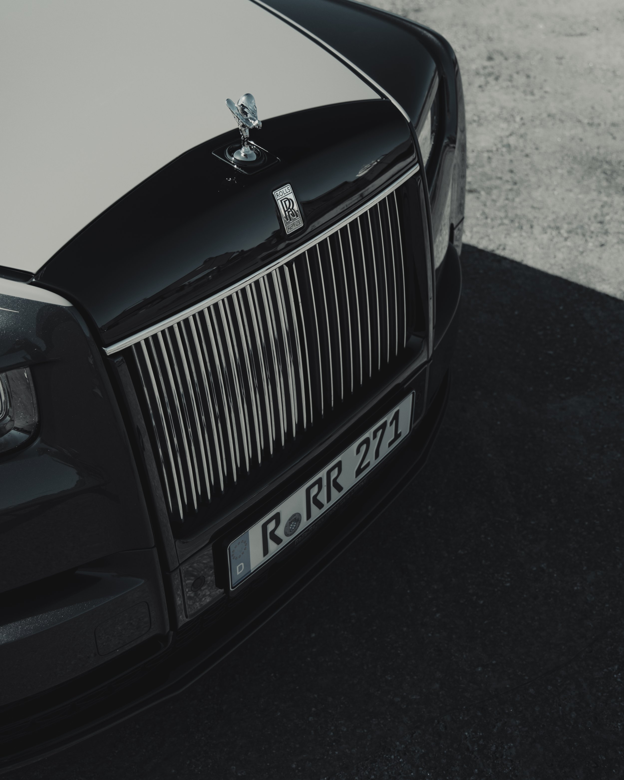 Close-up of a black Rolls-Royce car with a chrome grille and the Spirit of Ecstasy hood ornament, seen from the front and slightly above, on a paved surface.