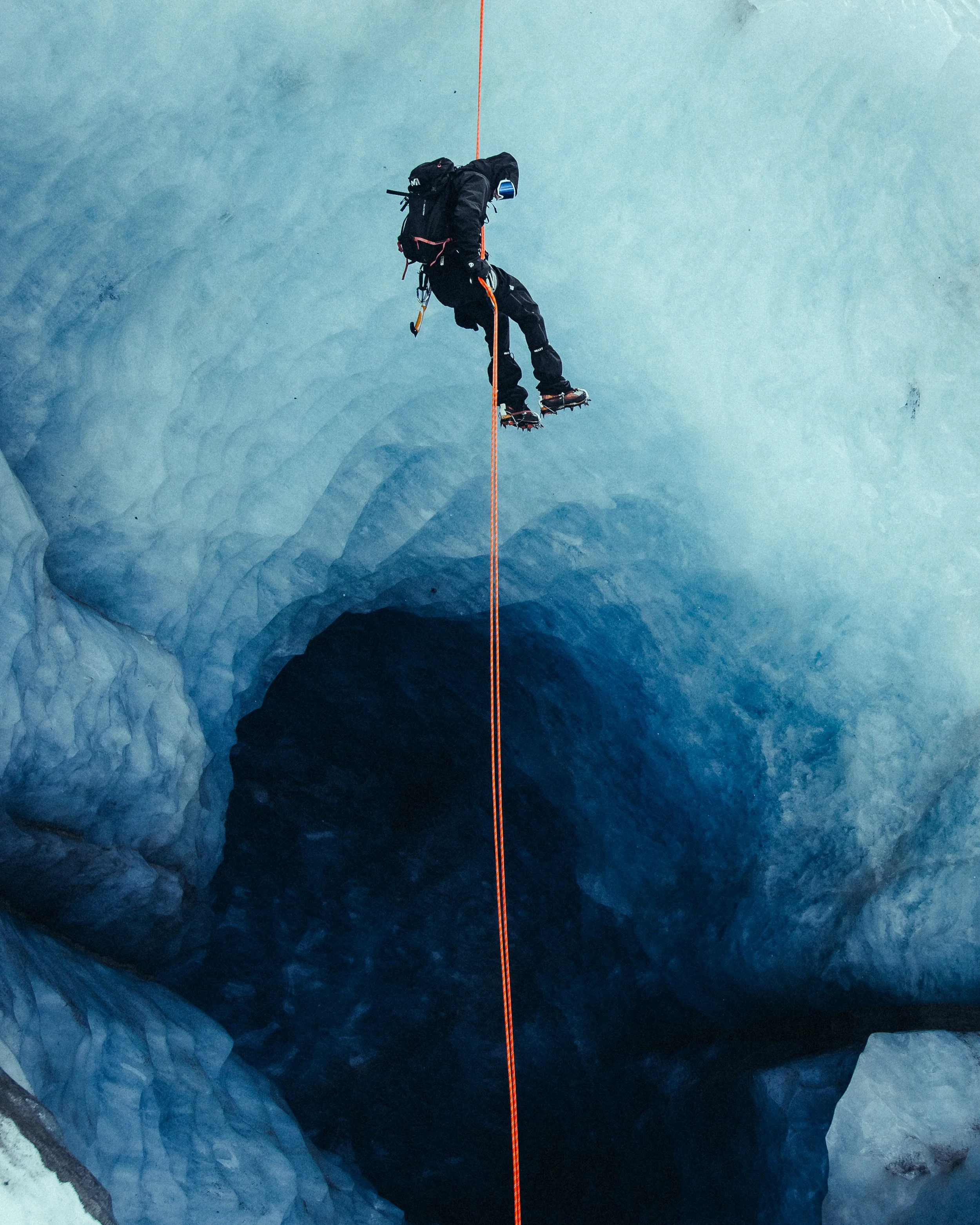 A person with climbing gear walking across a glacier using crampons, connected to an orange safety rope that extends into a dark ice crevasse below.