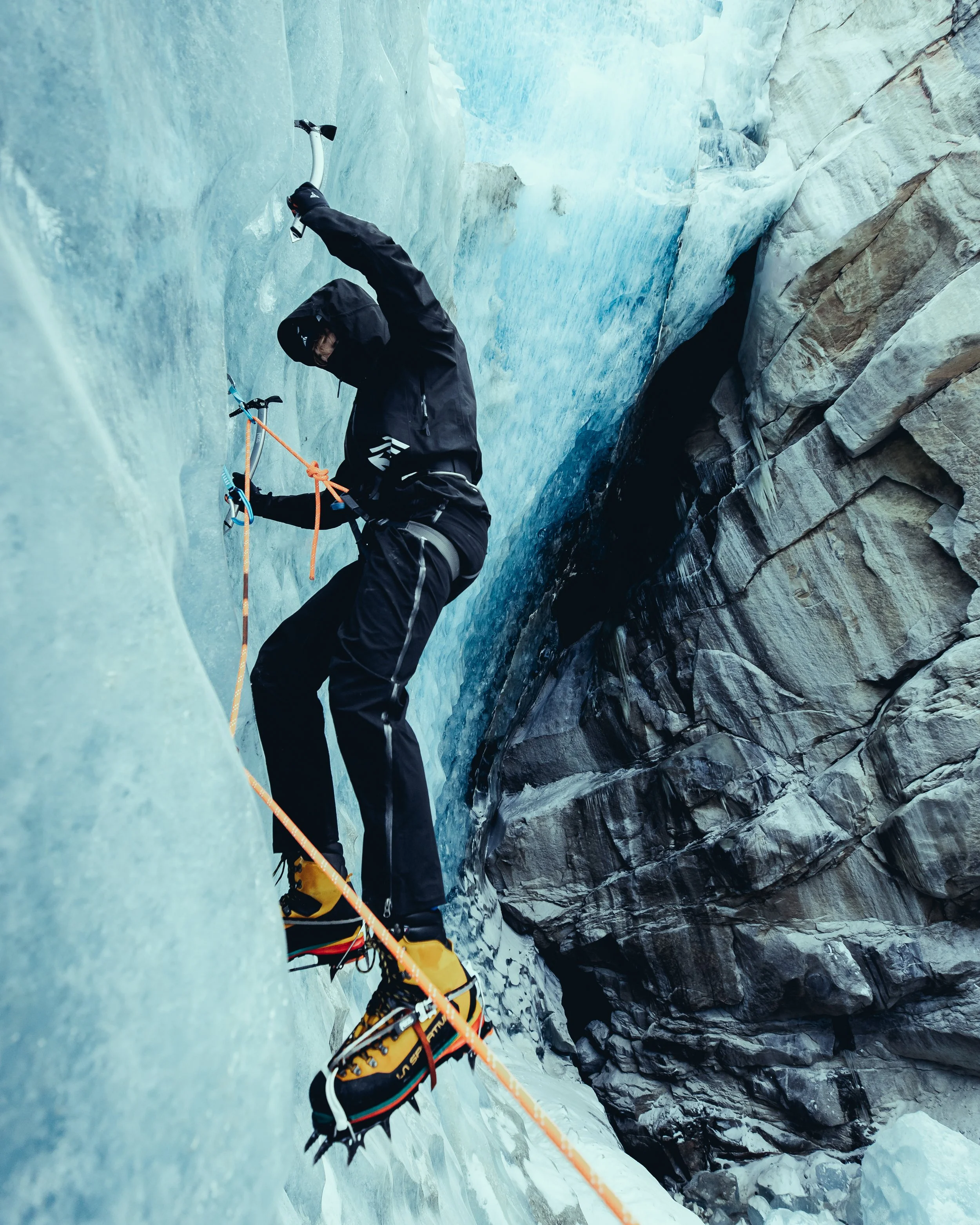 A person ice climbing on a frozen ice wall with rock formations visible to the right.