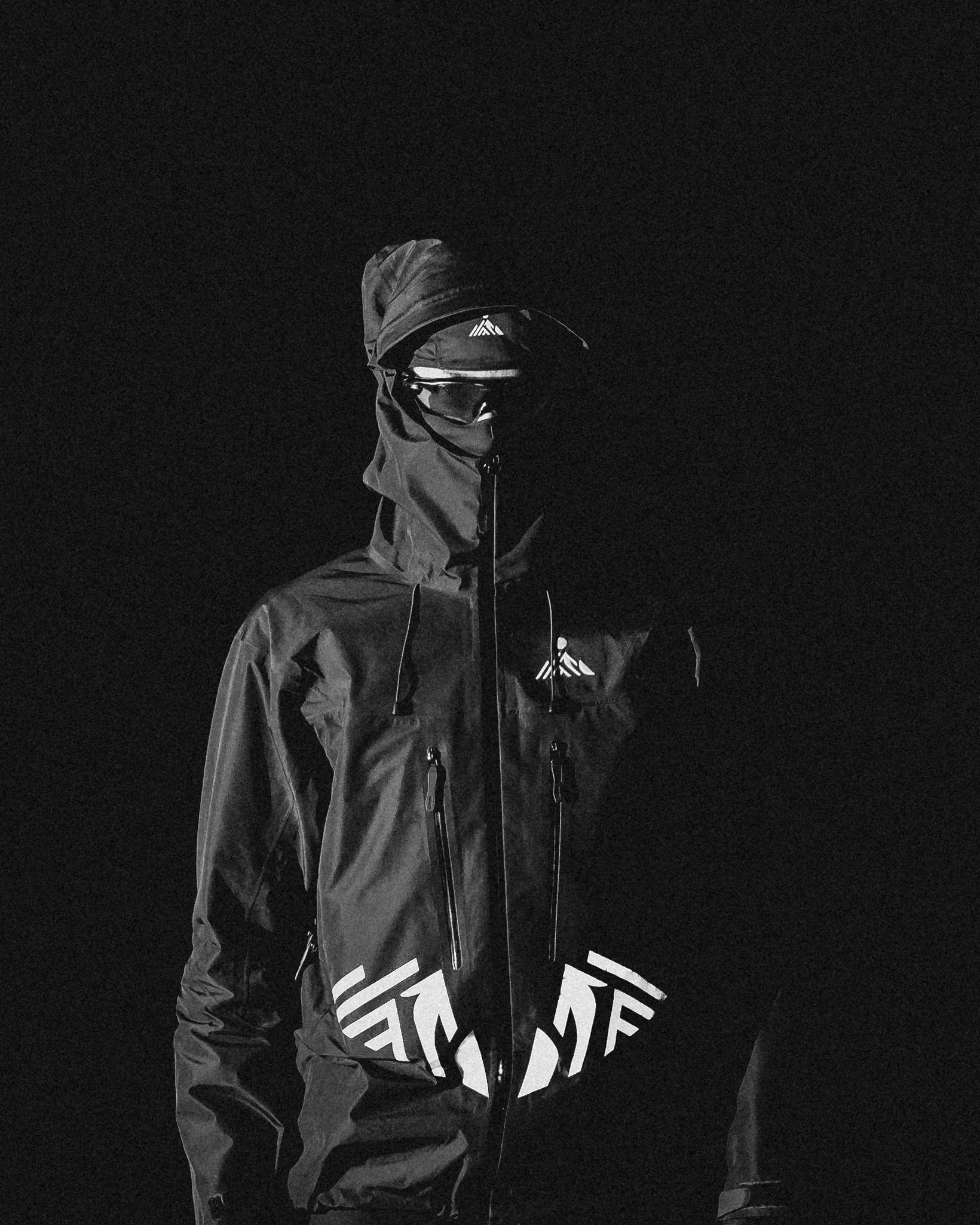 A person dressed in black outdoor gear, including a hooded jacket, goggles, and a hat, standing in darkness.
